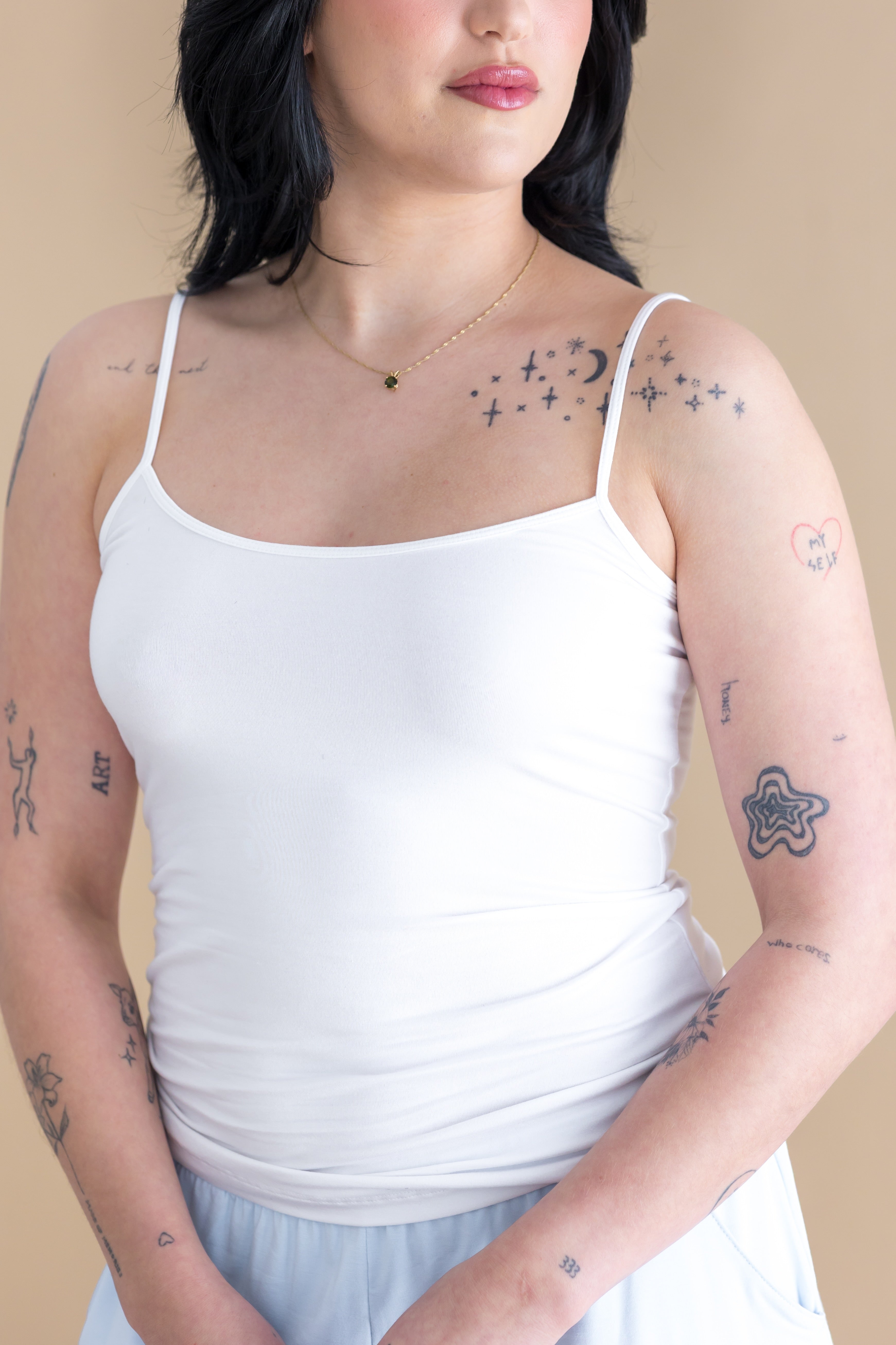 Woman wearing a white tank top with visible tattoos on her arms and neck against a beige background