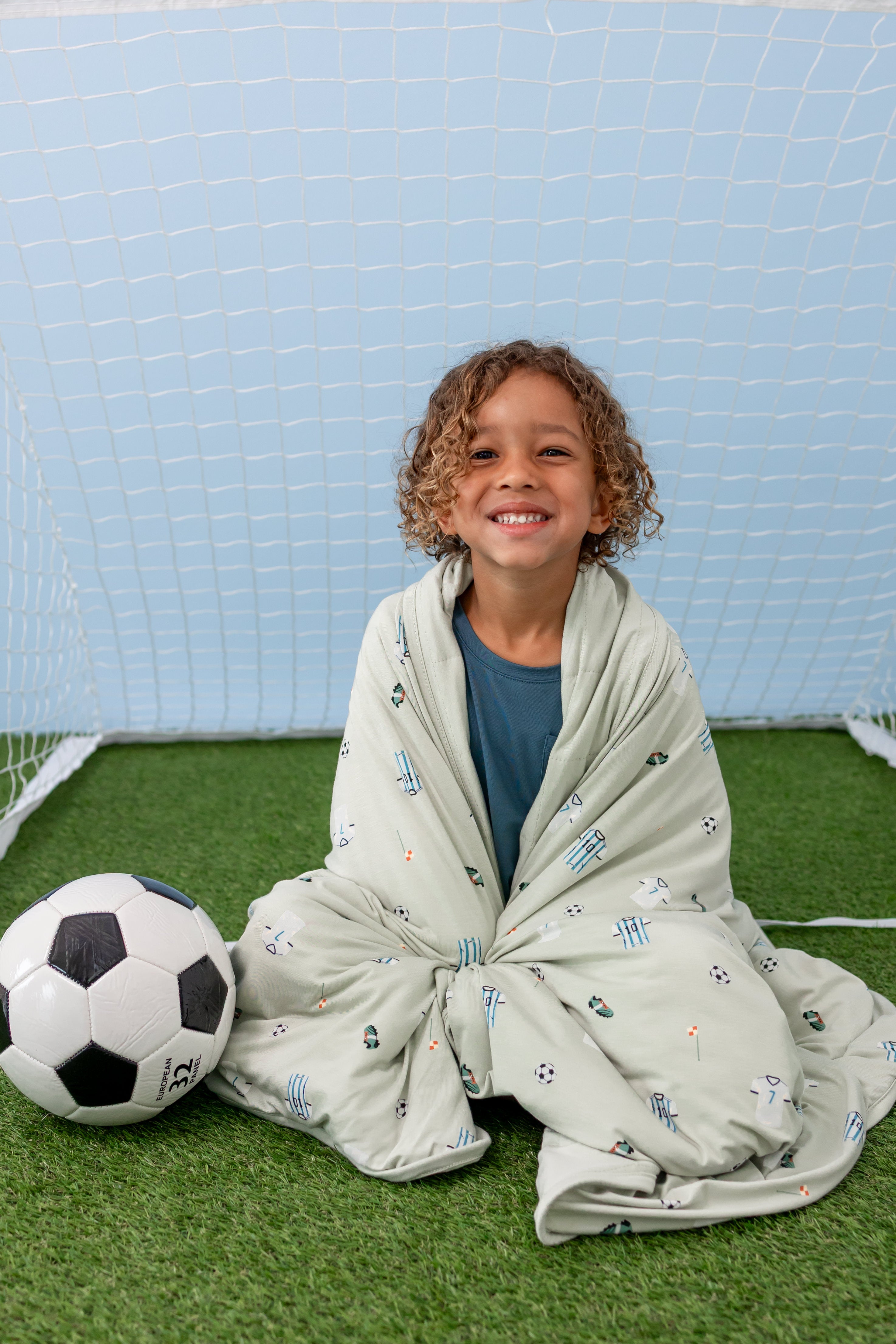 Child wrapped in Soccer print Toddler Blanket sitting in front of football post with a soccer ball on green grass