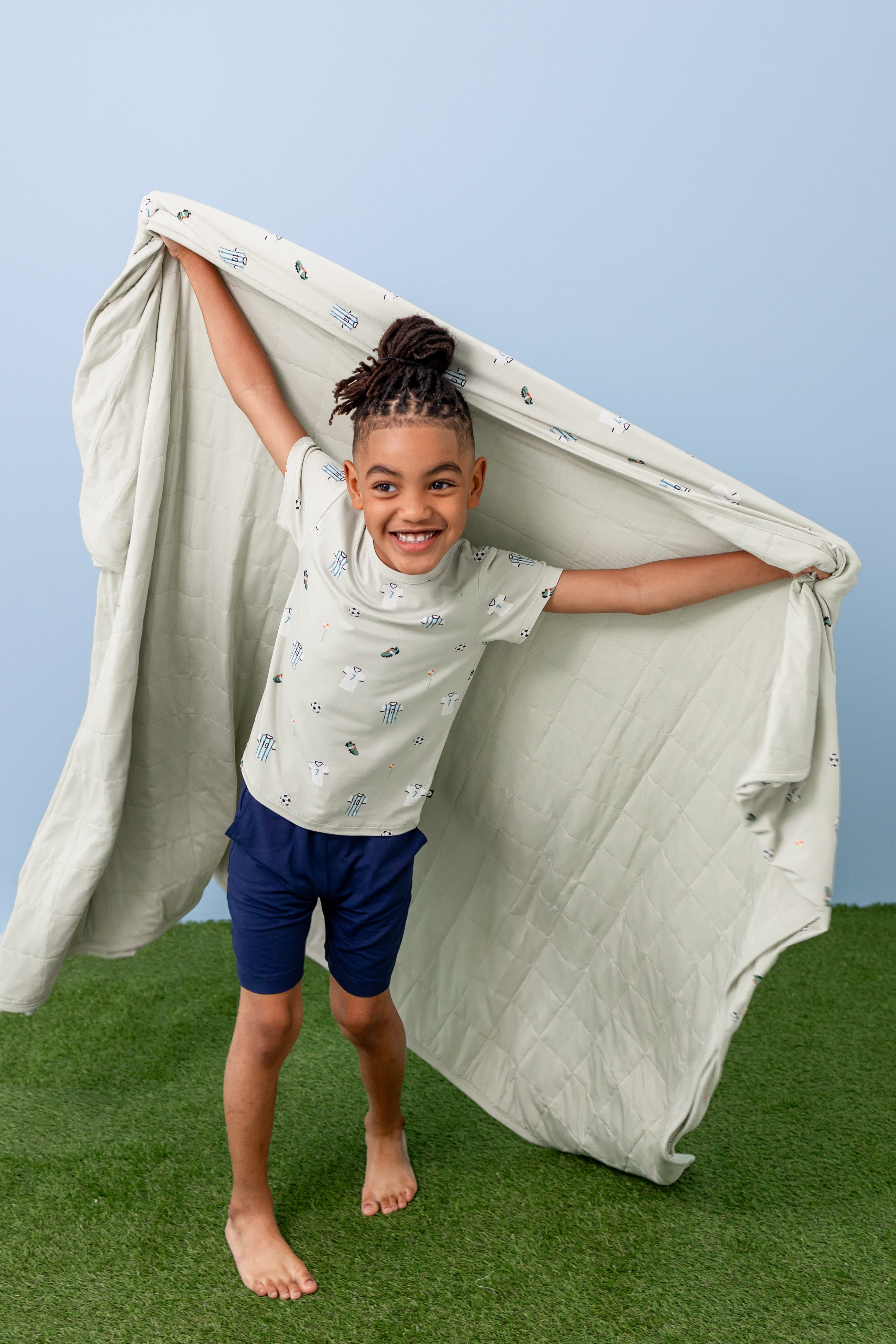 Child holding Youth blanket in Soccer print wearing a similar print Tee and navy shorts on a green grass