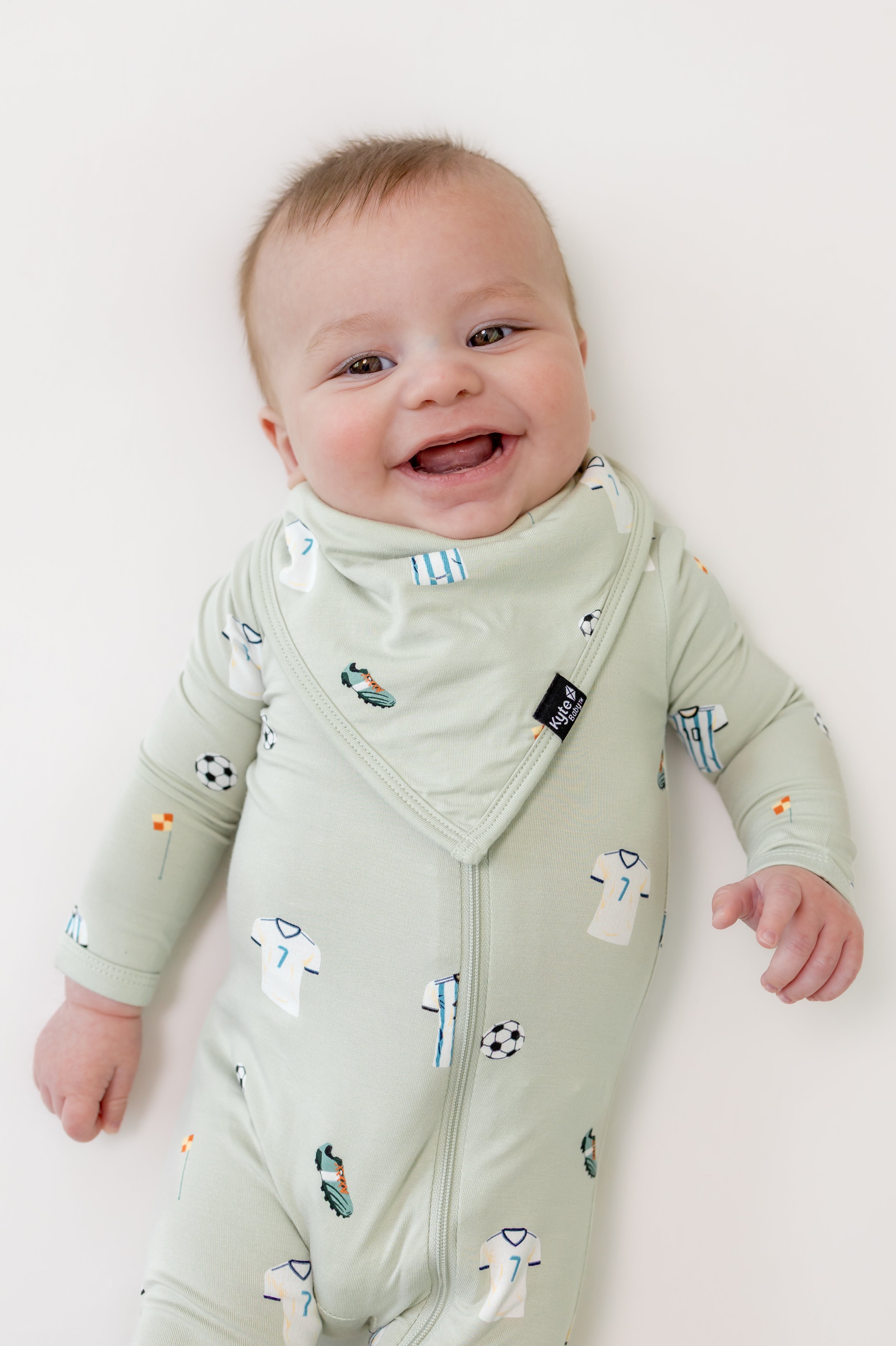 Baby wearing Soccer print Bib over a soccer print zippered romper on a beige background