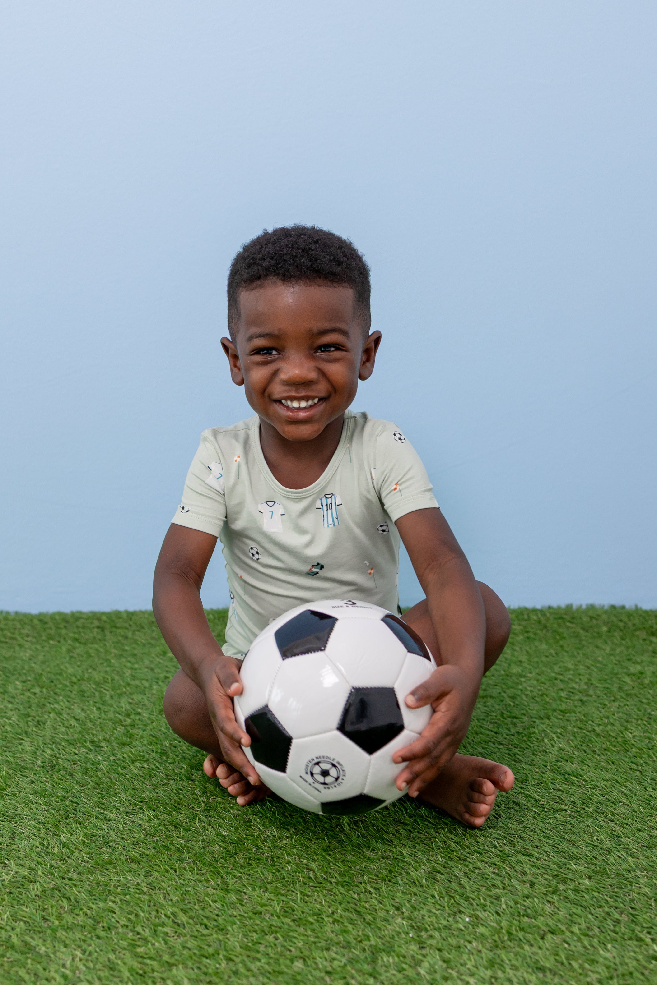 Child sitting with soccer ball in short sleeve pajama set in Soccer print on green grass
