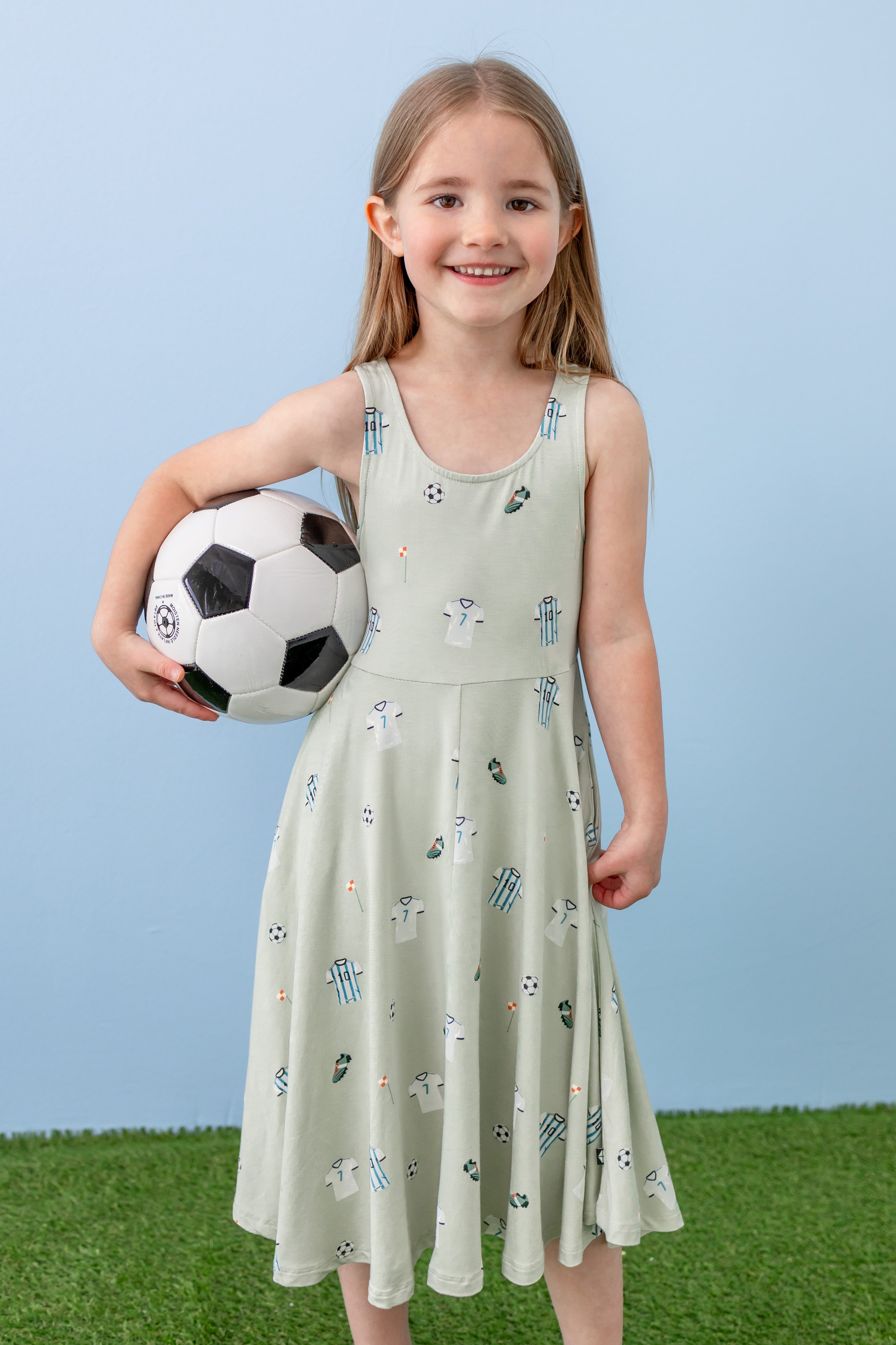 Child wearing Tank Twirl dress in Soccer print holding a soccer ball on green grass