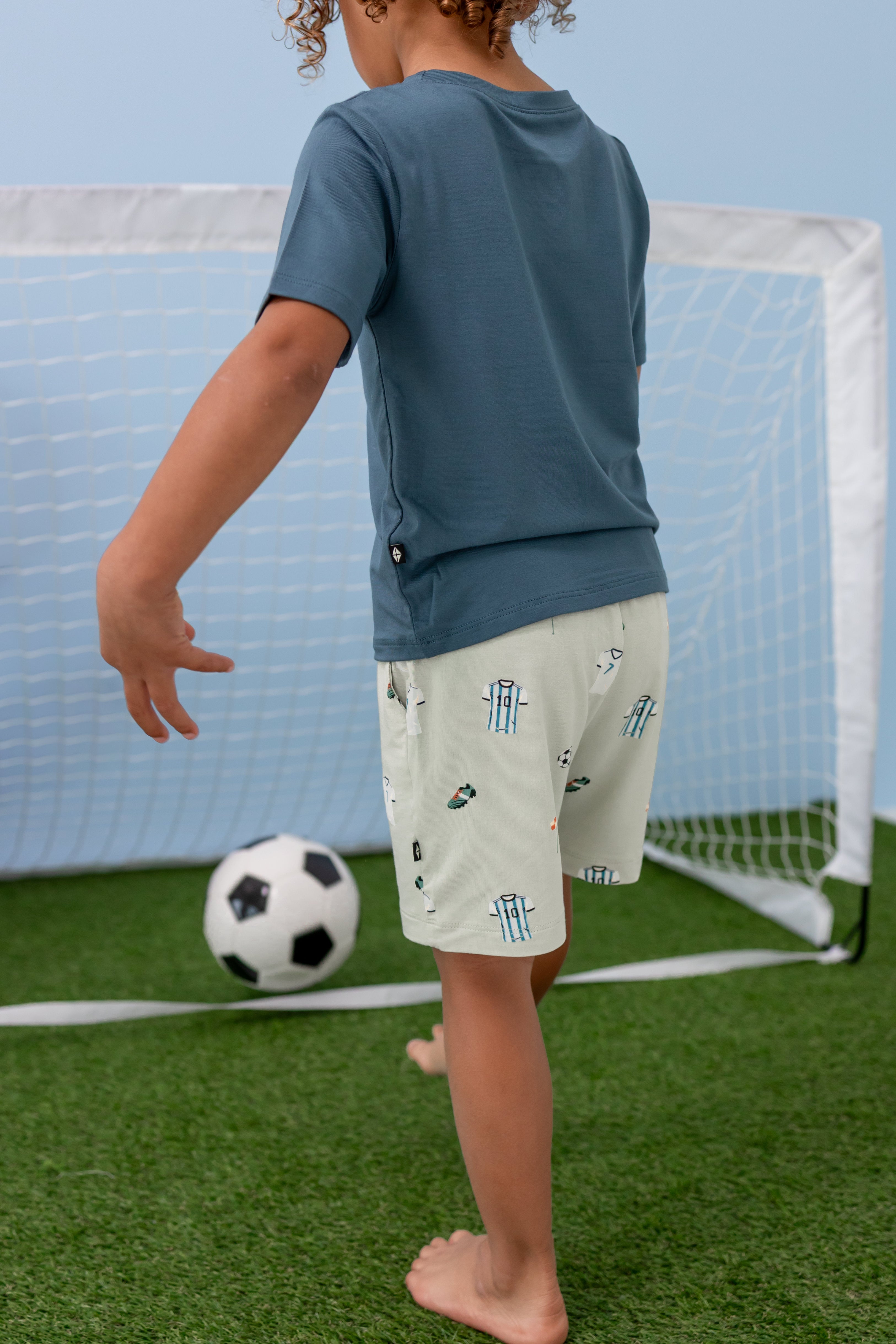 Toddler wearing blue tshirt and green soccer printed drawstring shorts kicking a soccer ball in a net