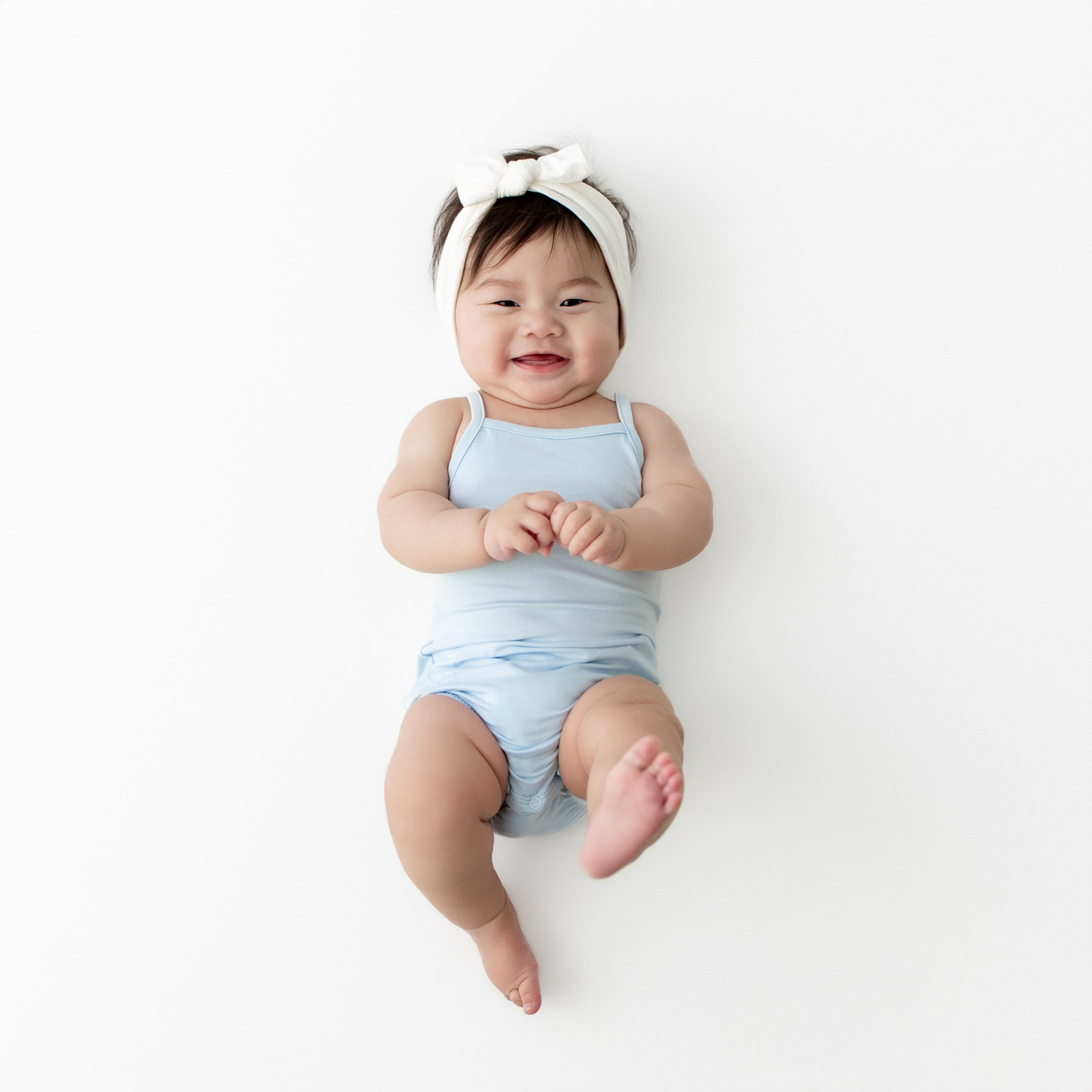 Young infant laying on a white surface wearing the Spaghetti Strap Leotard in Breeze with a white bow