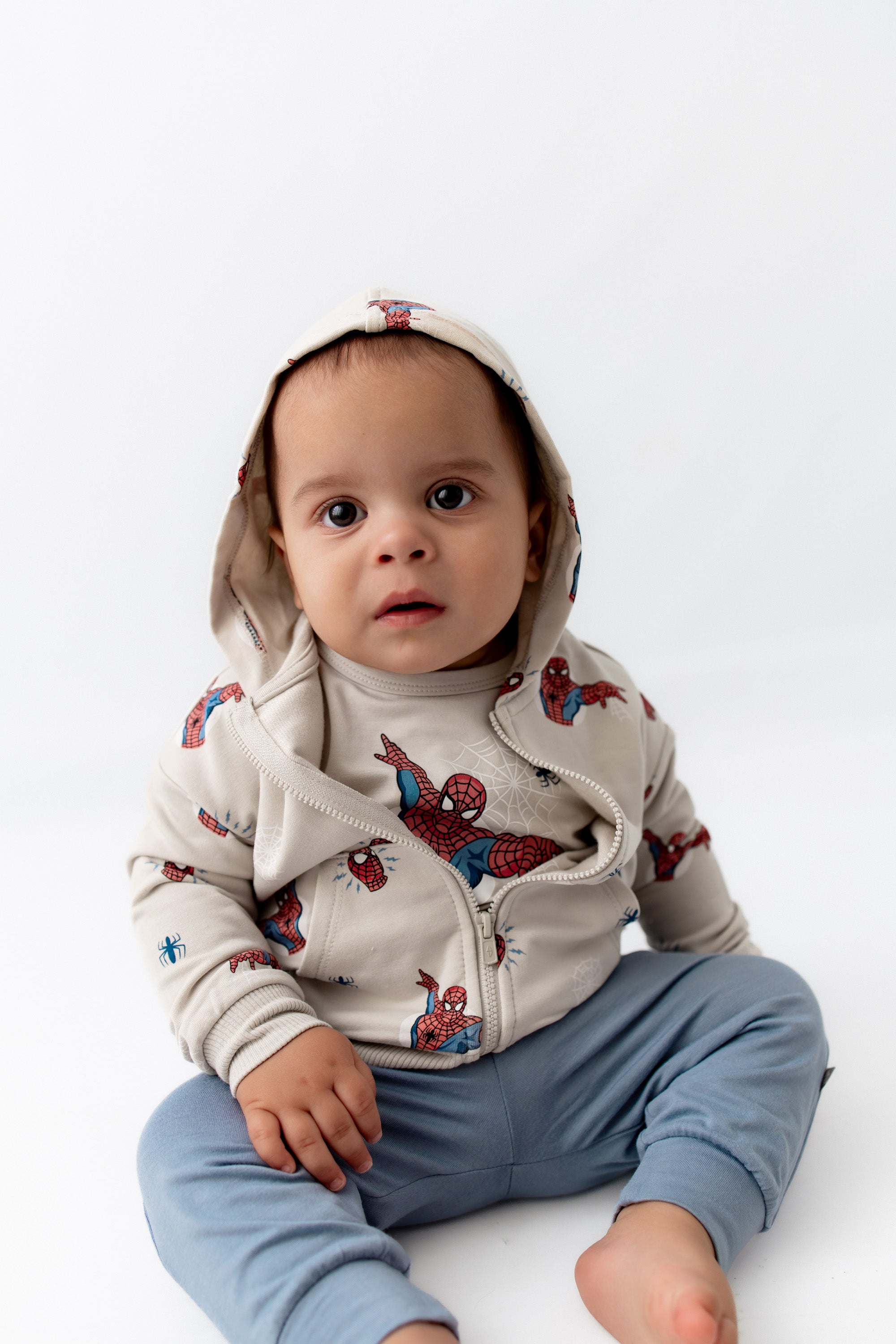 toddler sitting down wearing a spiderman jacket with the hoodie on his head.