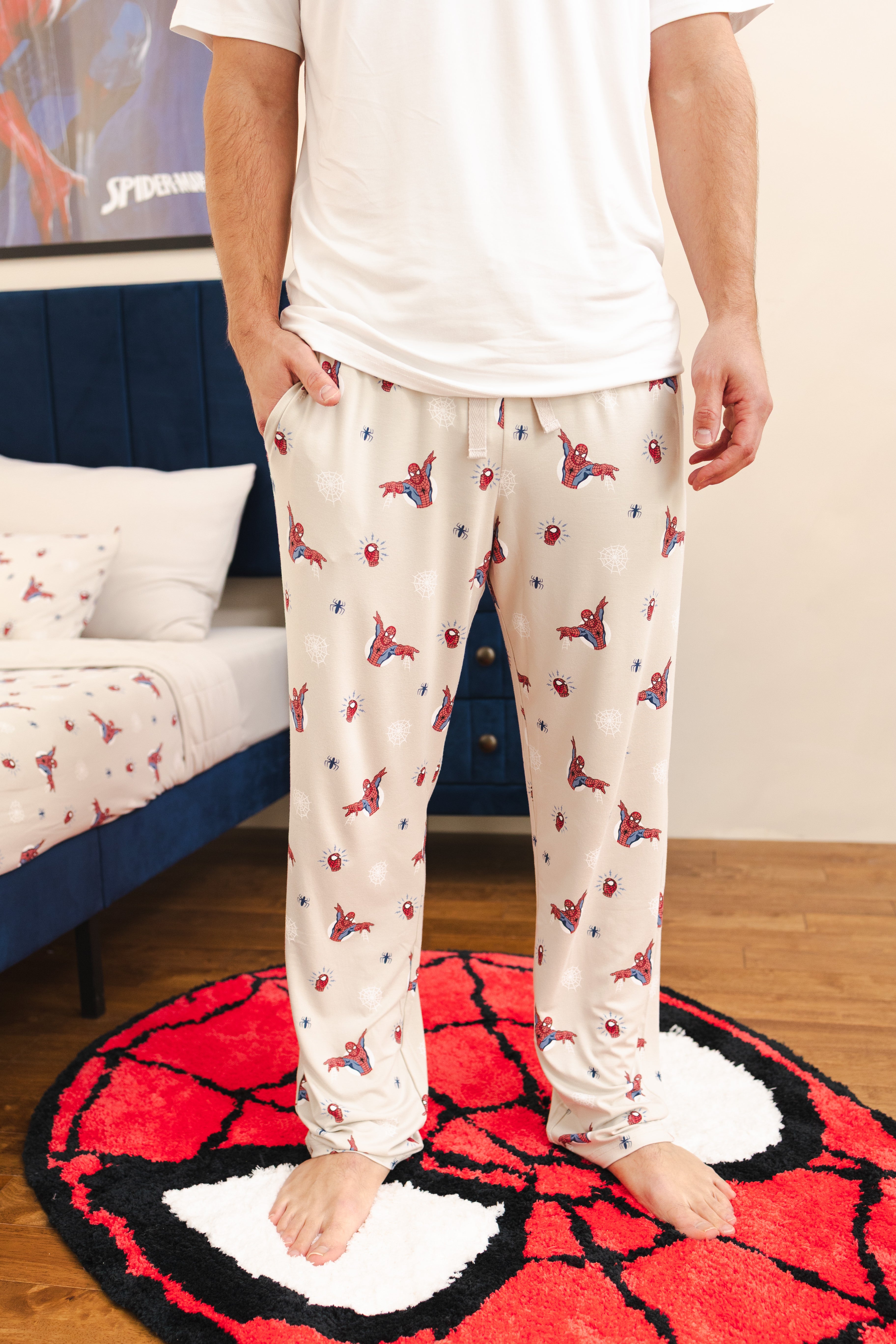A person wearing cream-colored pajama pants with a red and blue Spider-Man spider and web pattern, standing barefoot on a red and black Spider-Man themed rug in a bedroom setting.