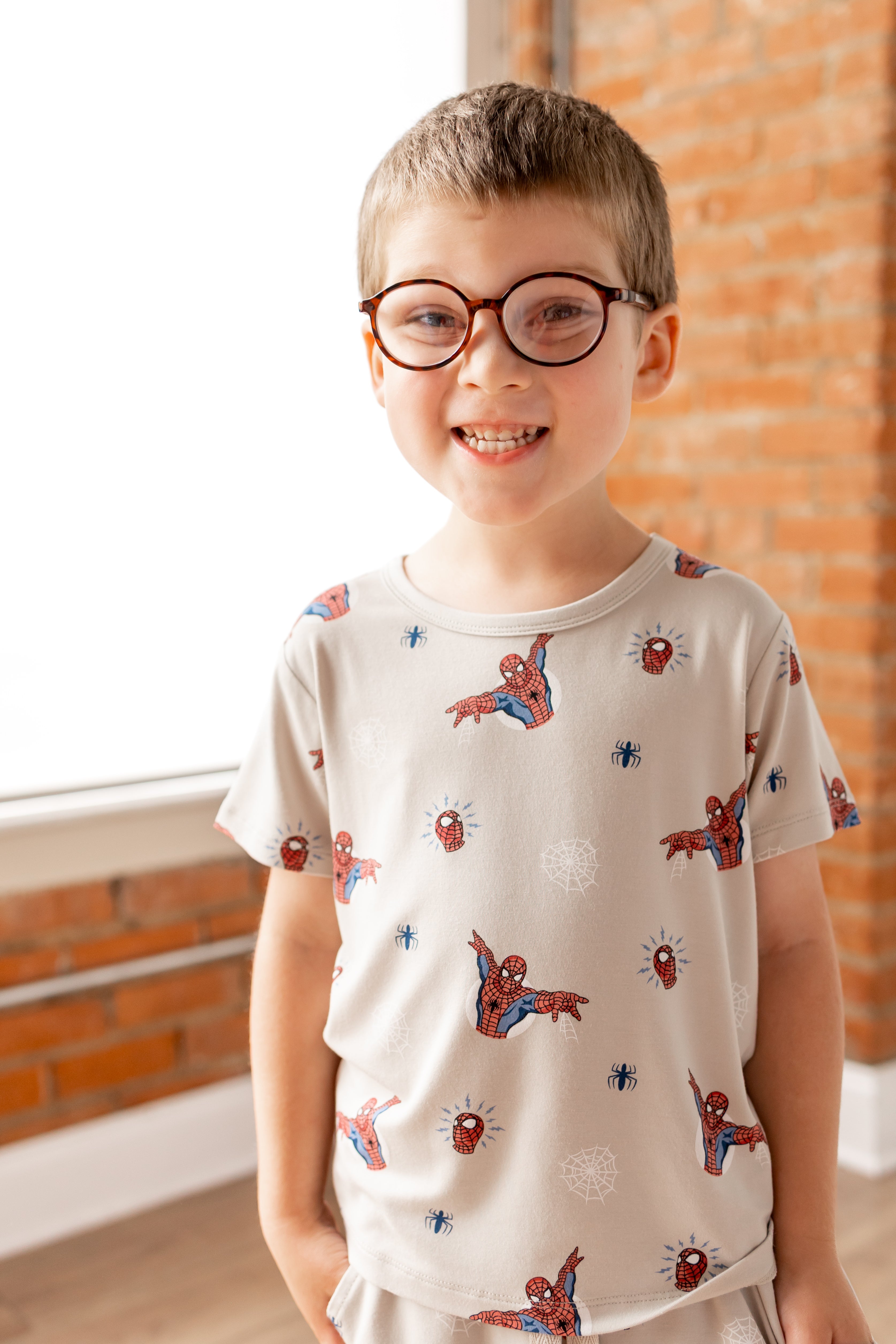 Boy in specs smiling in oat color Toddler Basic Tee with Spider-Man print in front of a brick wall