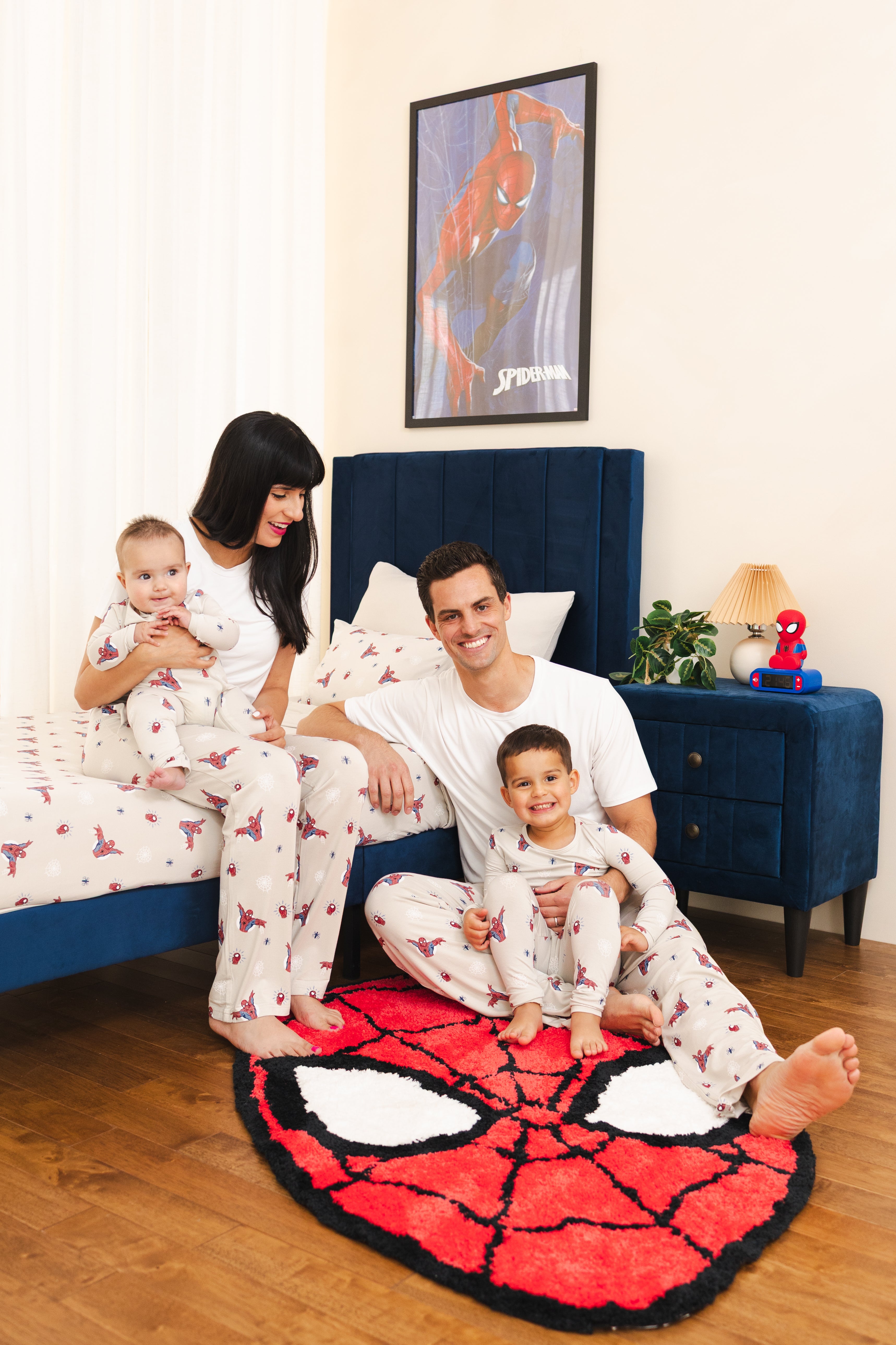A family of four wearing matching Spider-Man pajamas sits on a bed in a bedroom. The pajamas are white with a red and pink spider pattern.