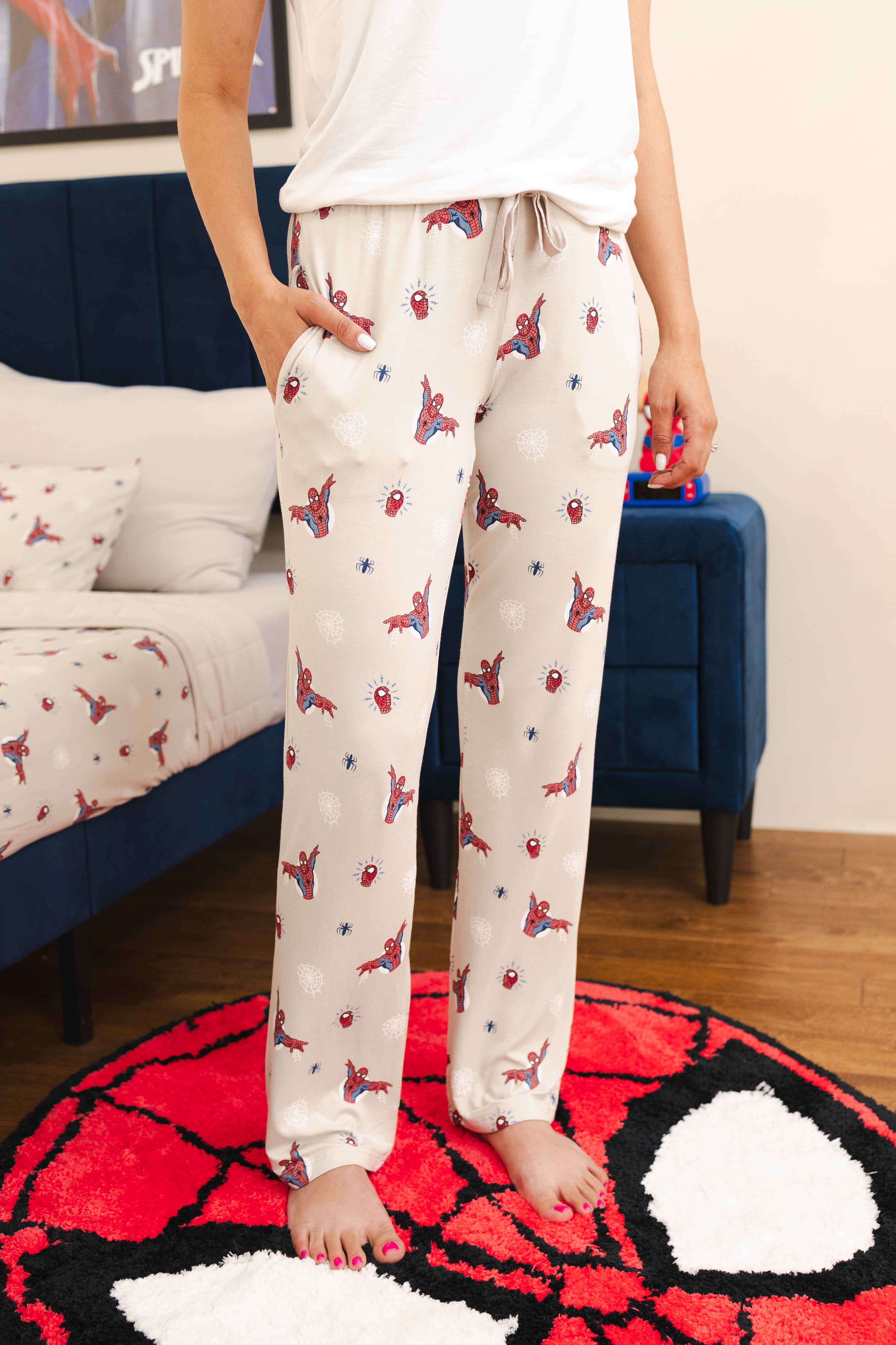 A person wearing cream pajama pants with an all-over Spider-Man print, standing barefoot on a red and white Spider-Man themed rug in a bedroom setting.
