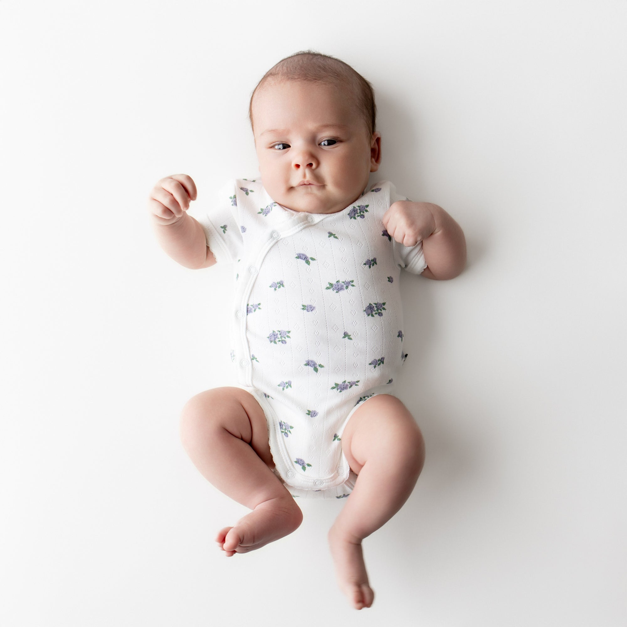 Newborn laying on a light neutral surface wearing the Pointelle Kimono Bodysuit in Spray Rose showing the picot trim