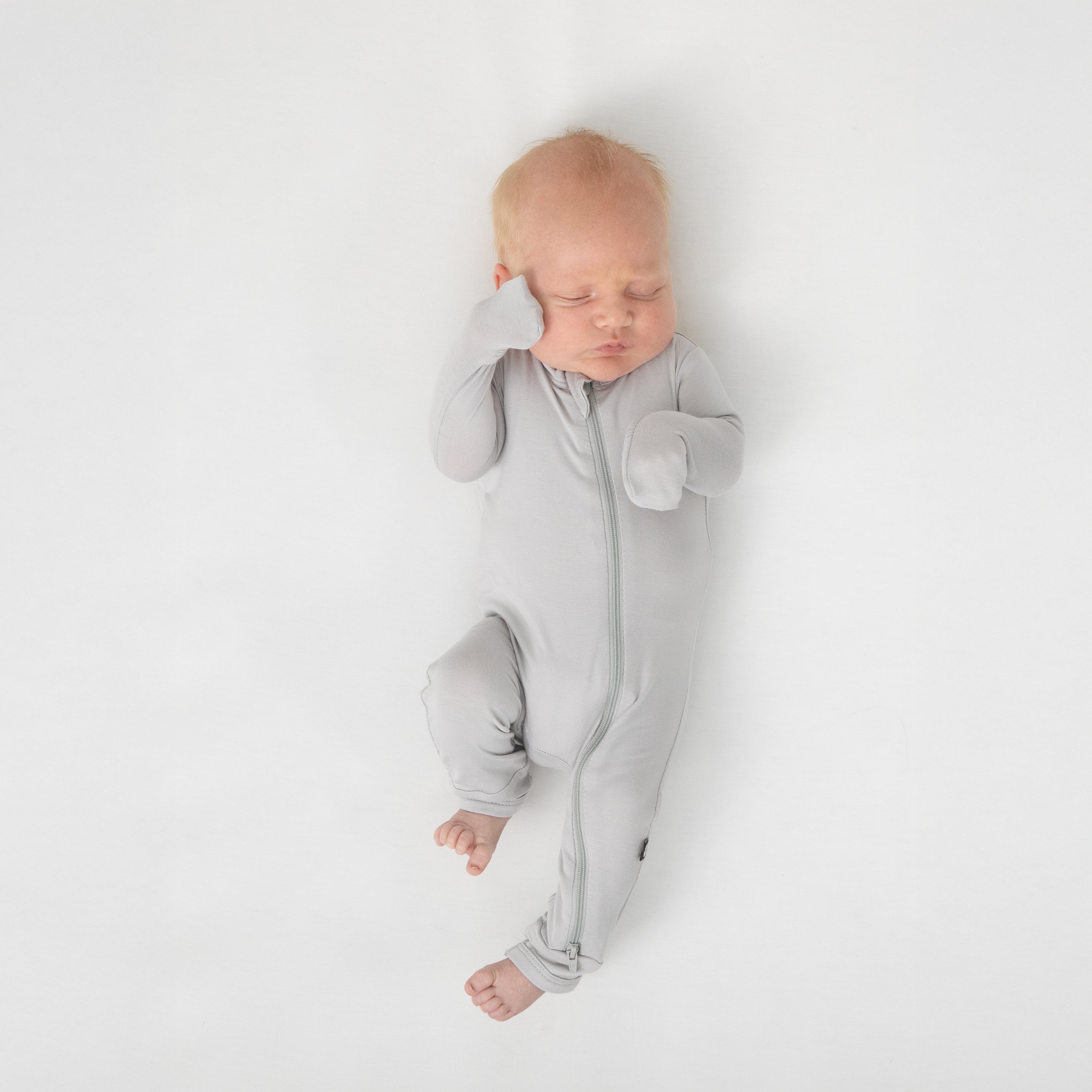 Sleeping newborn laying on a white crib sheet wearing the Zippered Footie in Storm