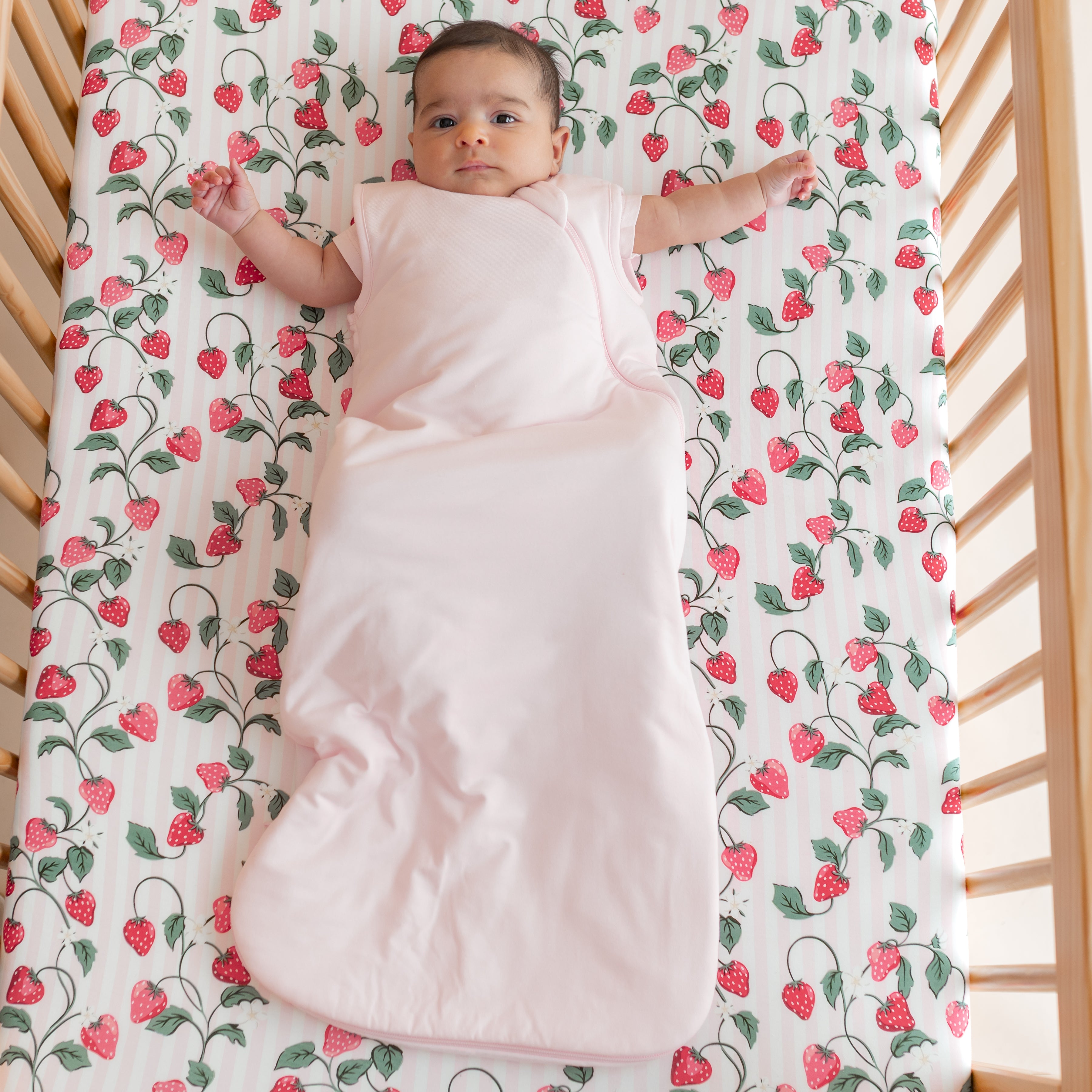 Infant laying in a crib on the breathable Crib Sheet in Strawberry Fields wearing a light pink sleep bag