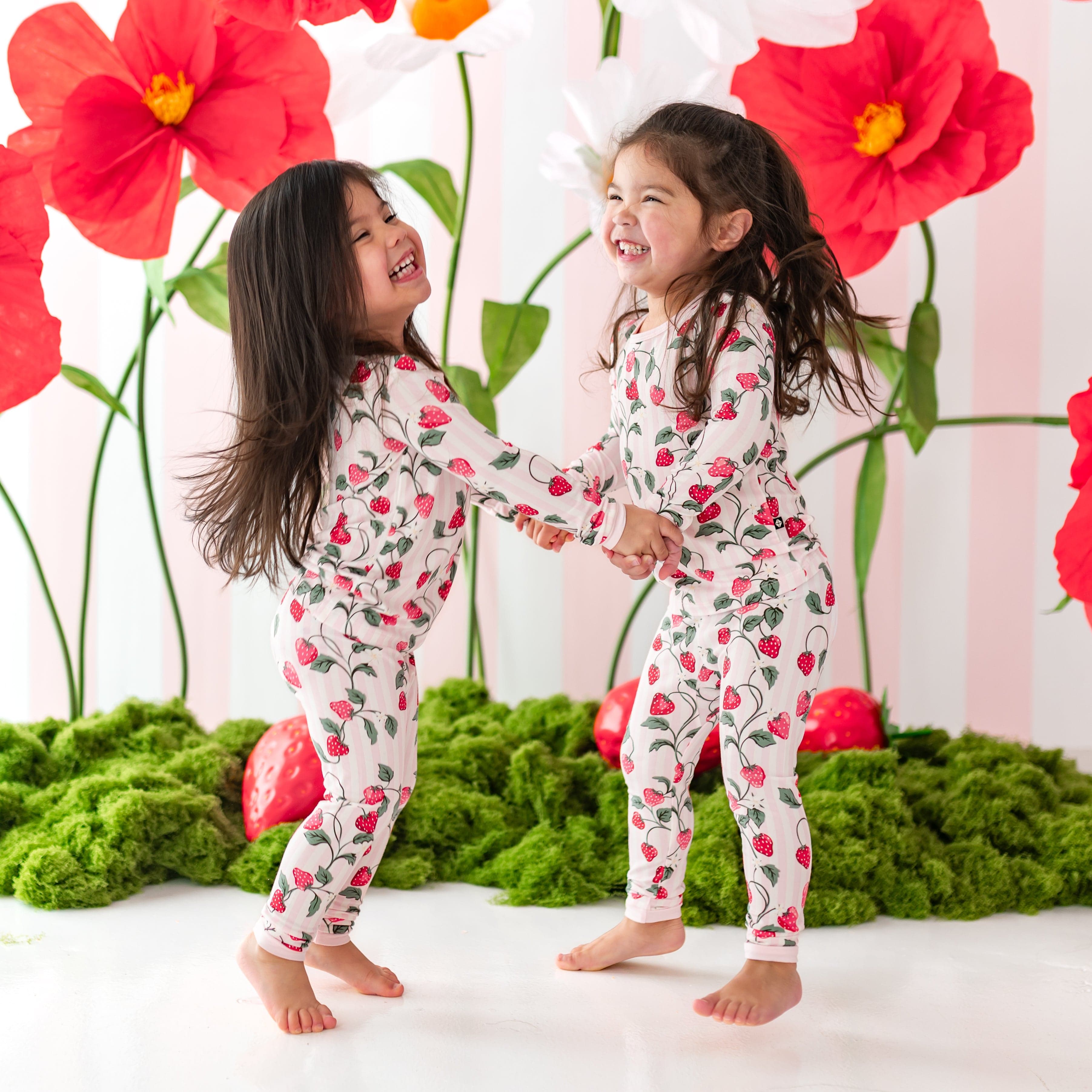 Two girls laughing, holding hands and jumping wearing the Long Sleeve Pajamas in Strawberry Fields in front of a strawberry backdrop