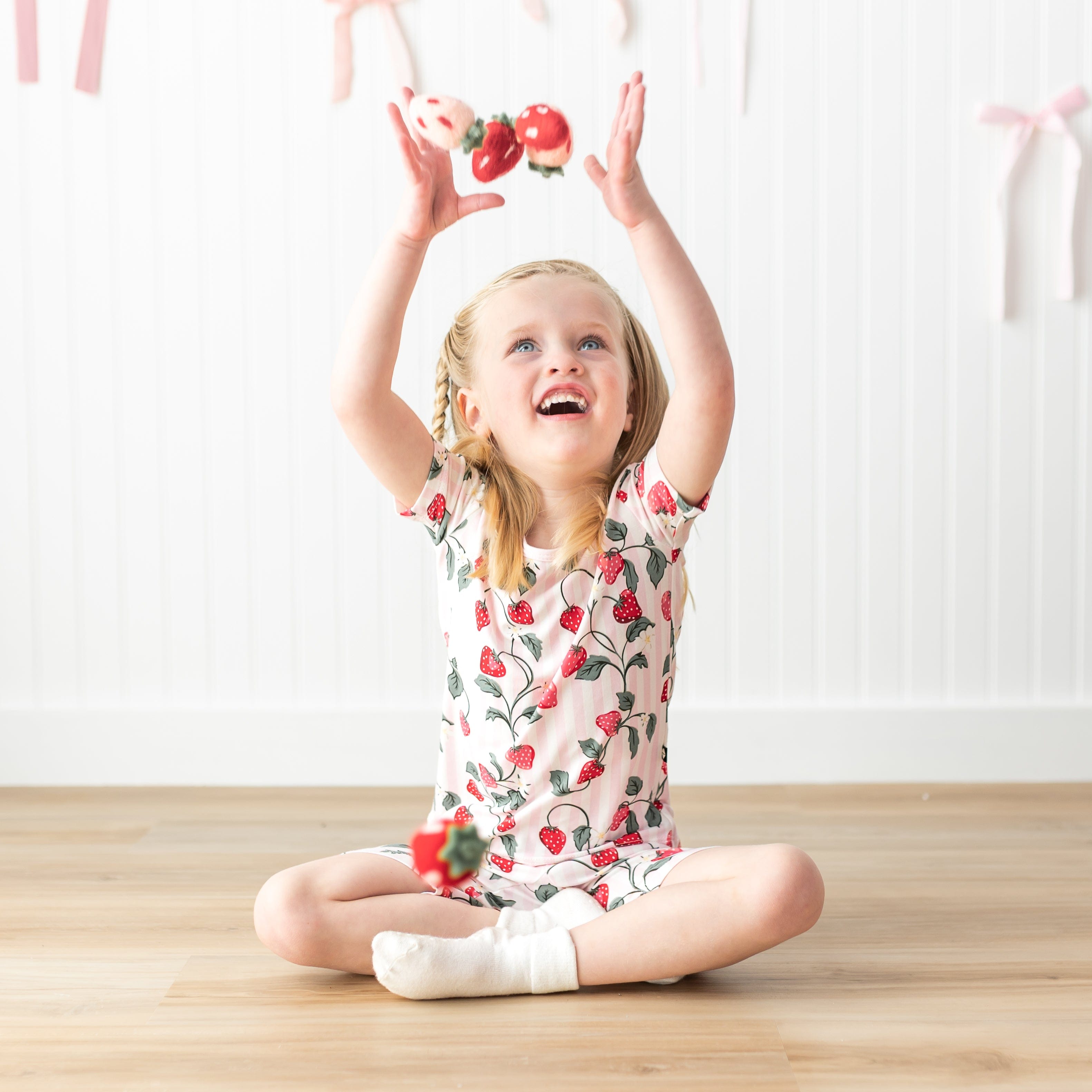 Young girl sitting cross legged on the floor wearing the Short Sleeve Pajamas in Strawberry Fields throwing plush toy strawberries in the air