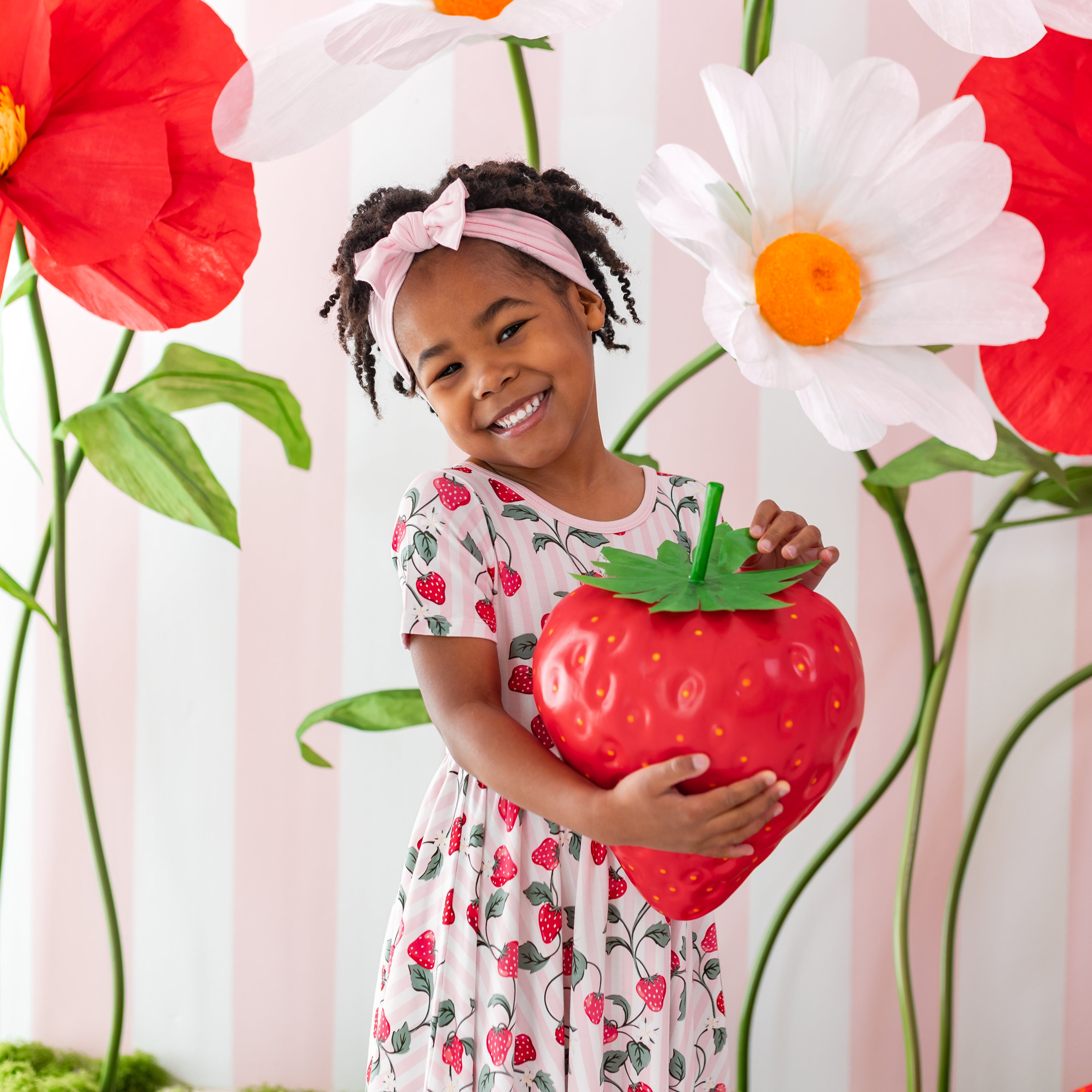 Young girl standing wearing the Twirl Dress in Strawberry Fields holding a prop strawberry