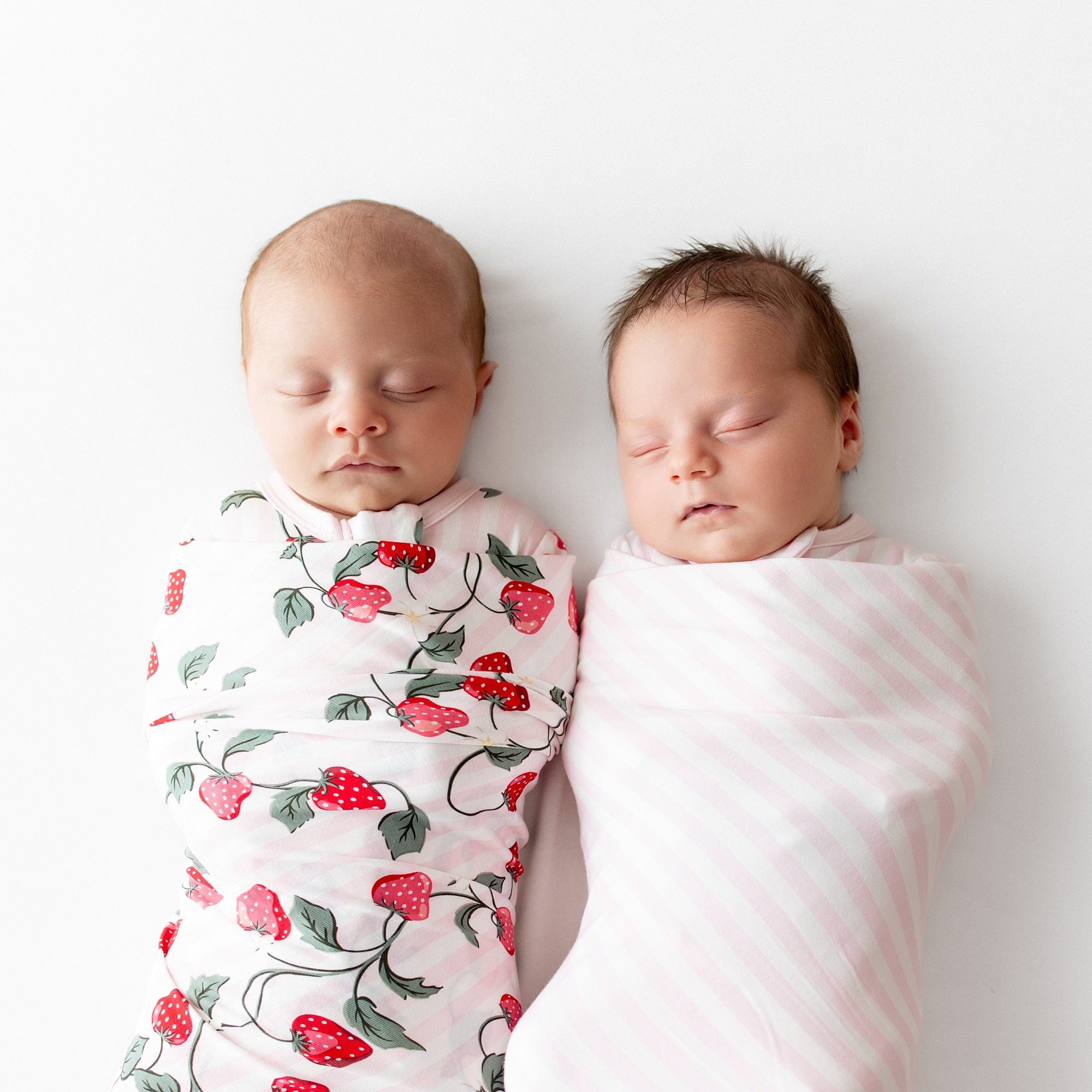 Two newborn laying side by side swaddled in the Swaddle Blanket in Strawberry Fields and Small Sakura Stripe