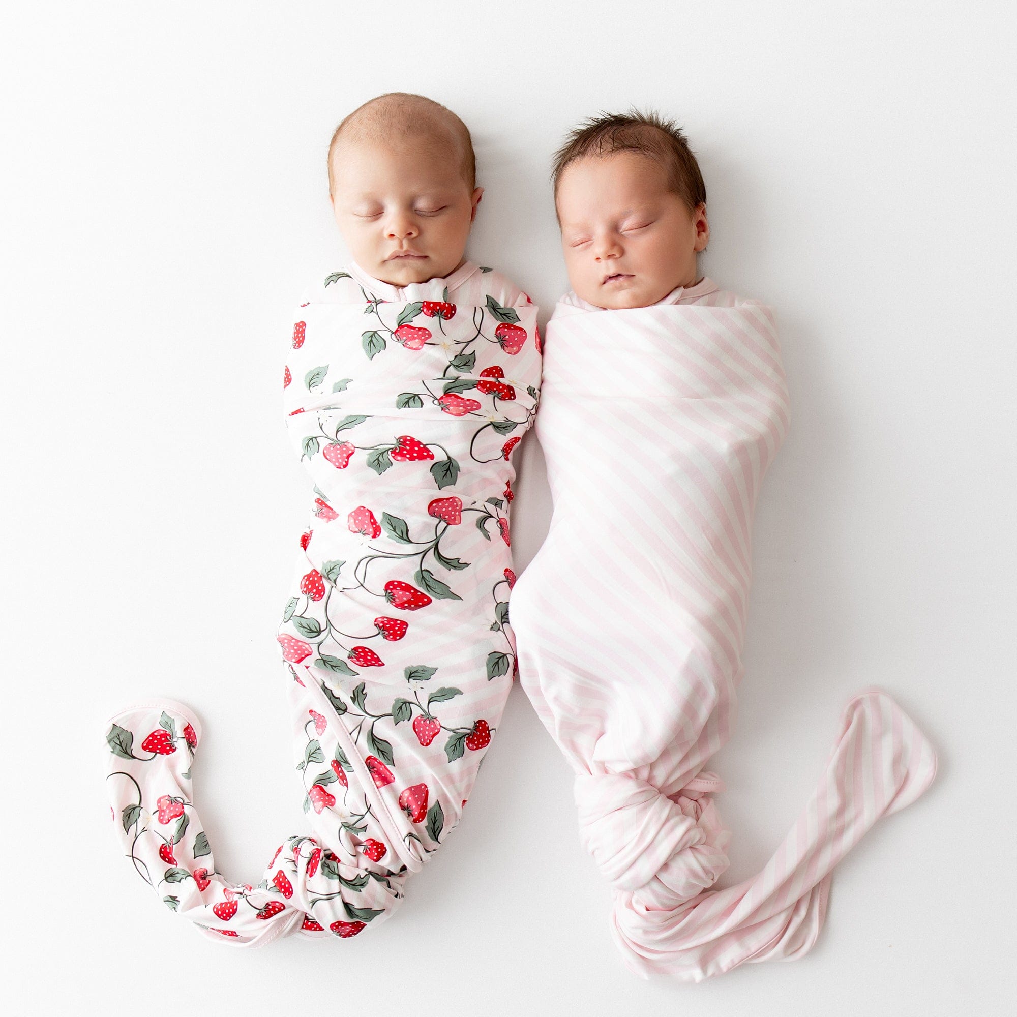 Two sleeping newborns laying side by side swaddled in the Strawberry Fields and Small Sakura Stripe Swaddle Blanket