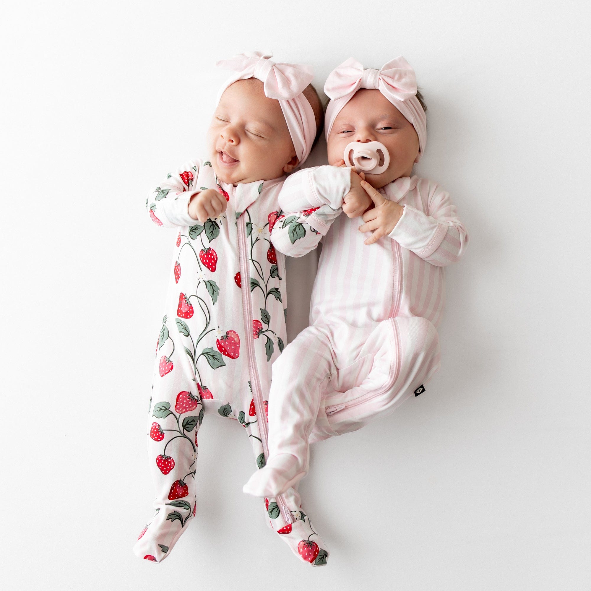 Two newborns laying side by side wearing the Zippered Footie in Small Sakura Stripe and Strawberry Fields with Sakura bow headbands