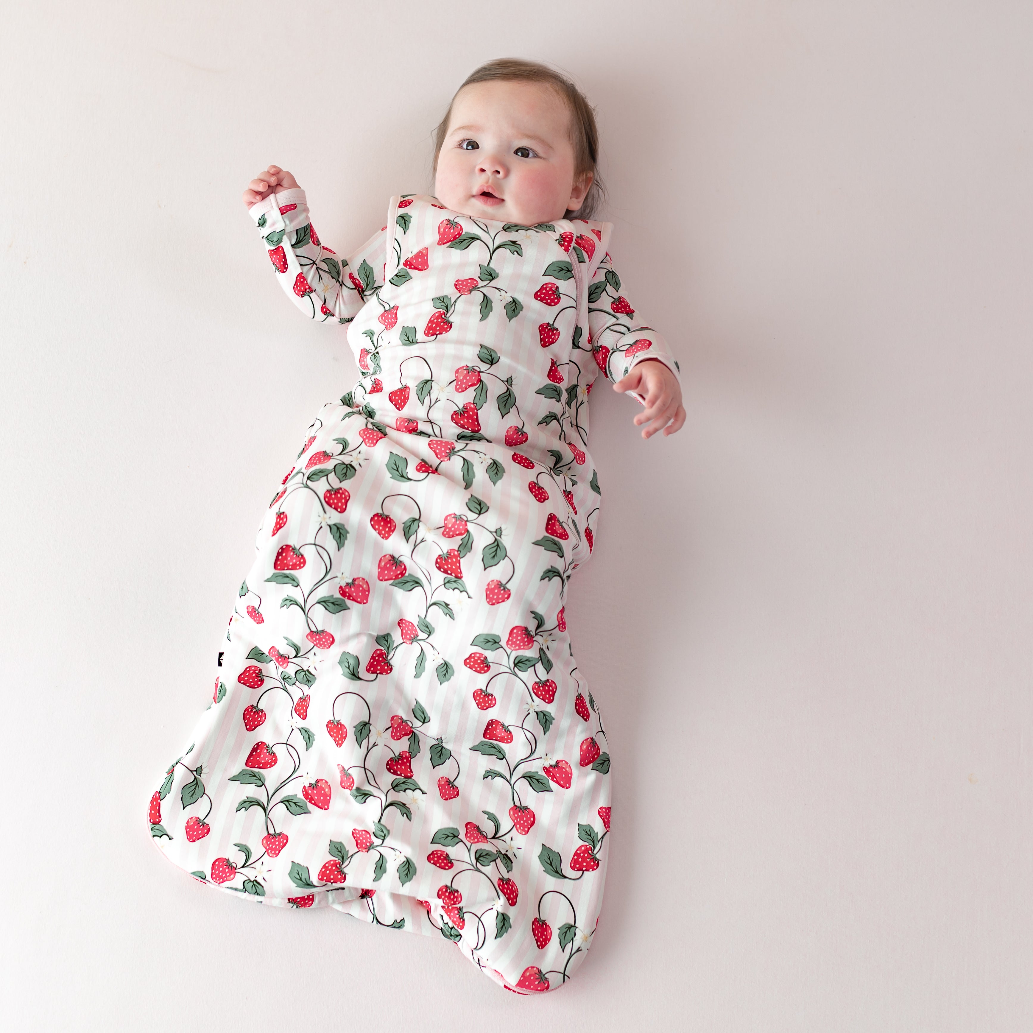 Young infant laying on a light pink blanket wearing the Sleep Bag in Strawberry Fields 1.0 with matching romper on underneath
