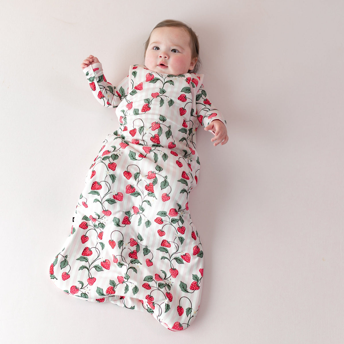 Young infant laying on a light pink blanket wearing the Sleep Bag in Strawberry Fields 1.0 with matching romper on underneath