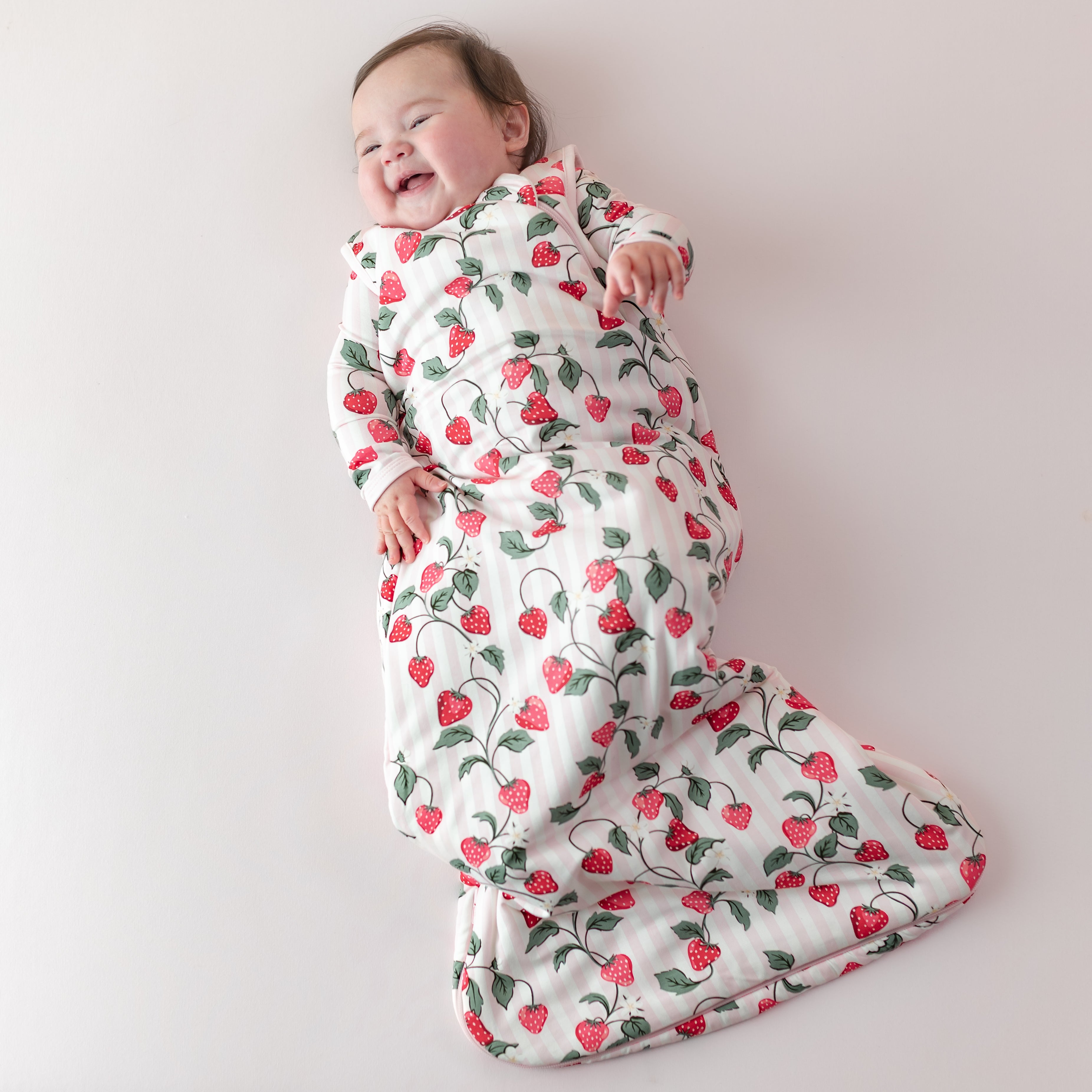 Young infant smiling laying on a light pink surface wearing the breathable Sleep Bag in Strawberry Fields 1.0 with a matching romper on underneath