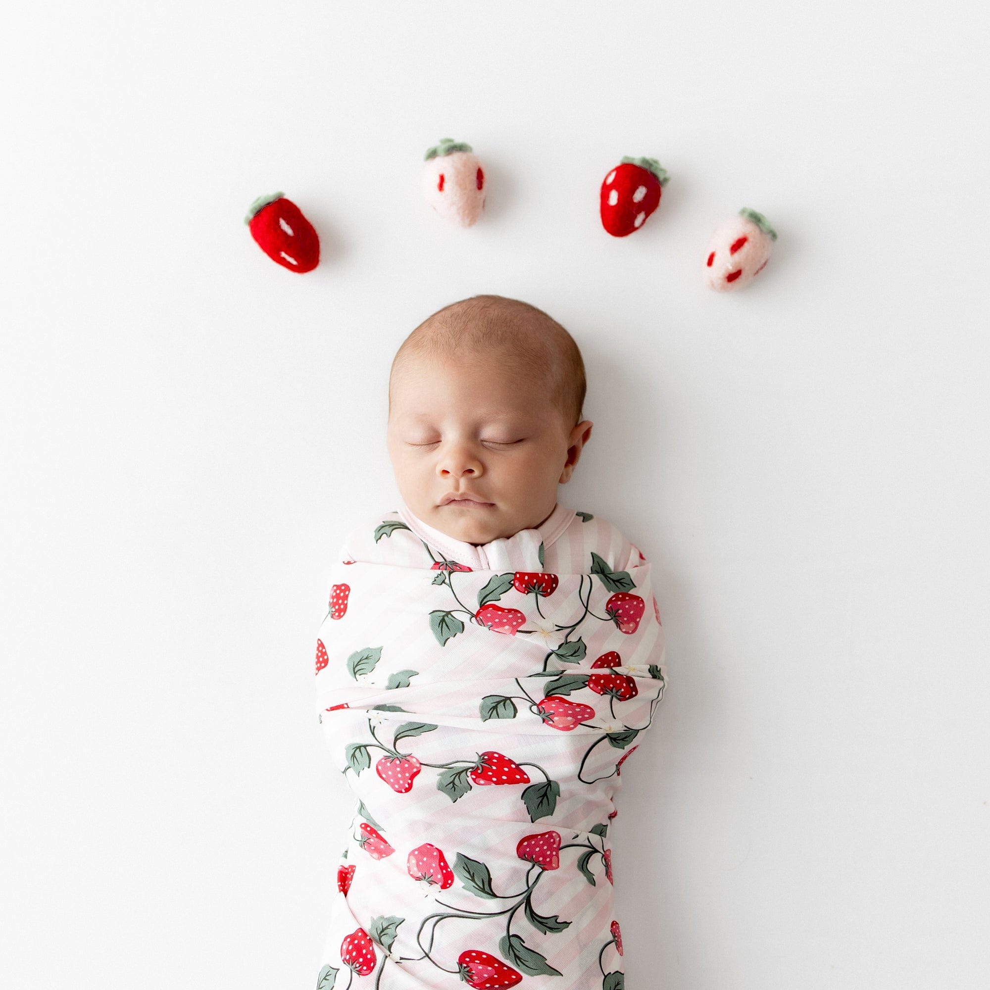 Close up of newborn swaddled in the Swaddle Blanket in Strawberry Fields with plush toy strawberries above her head