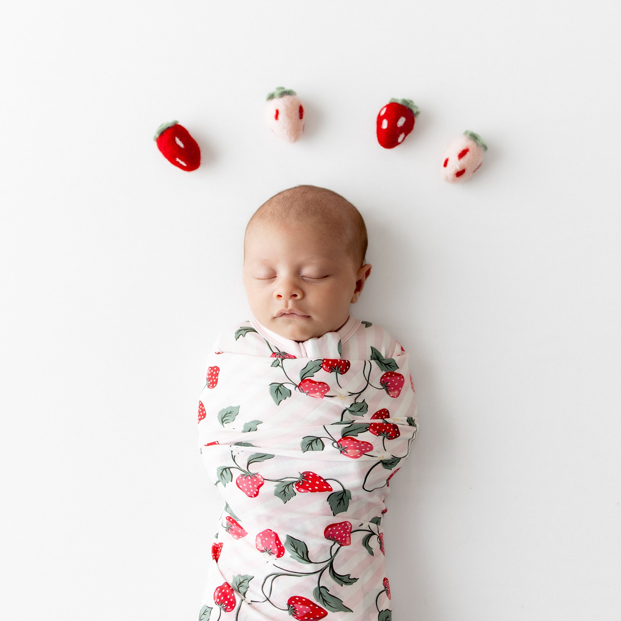 Close up of newborn swaddled in the Swaddle Blanket in Strawberry Fields with plush toy strawberries above her head