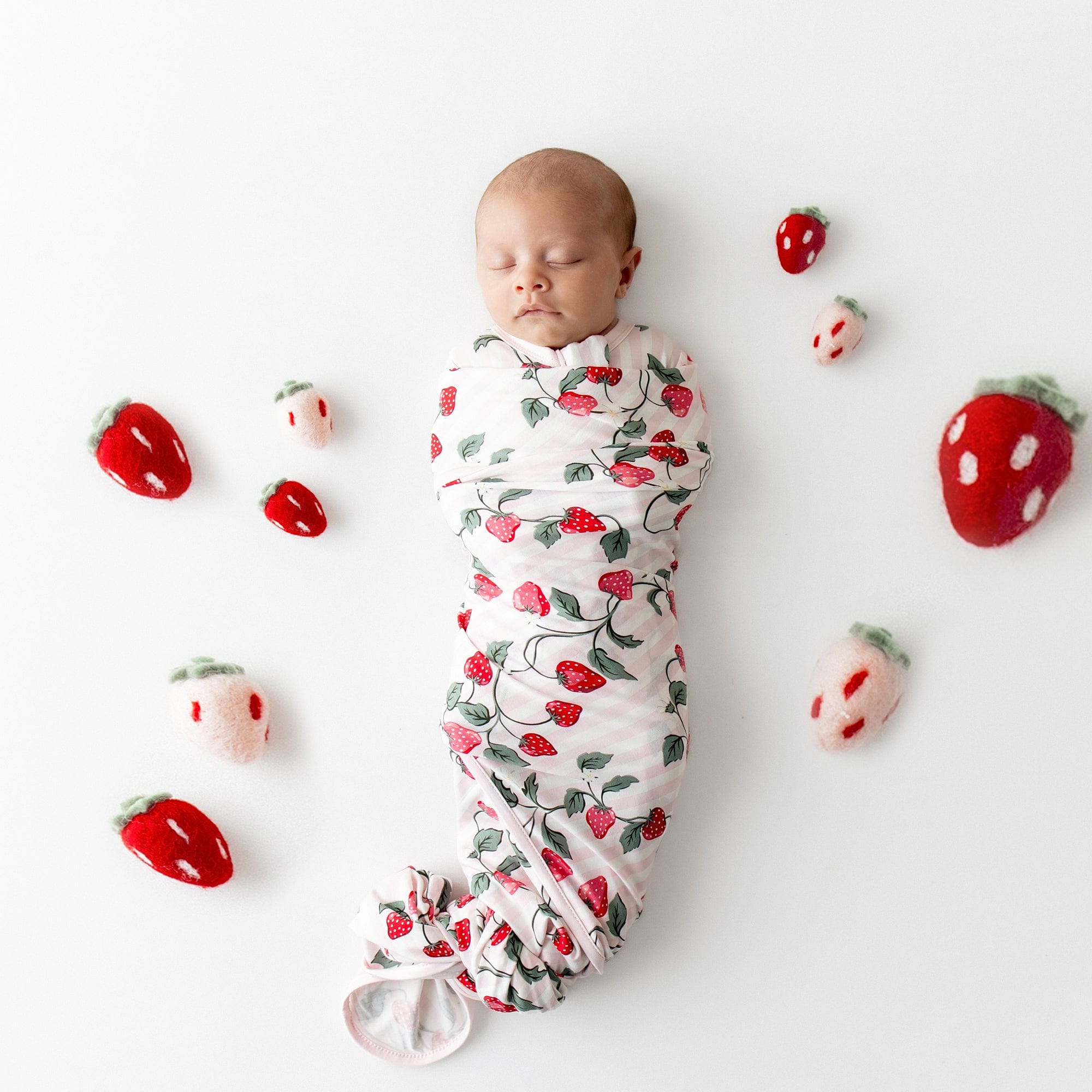 Sleeping newborn swaddled in the Swaddle Blanket in Strawberry Fields surrounded by plush toy strawberries