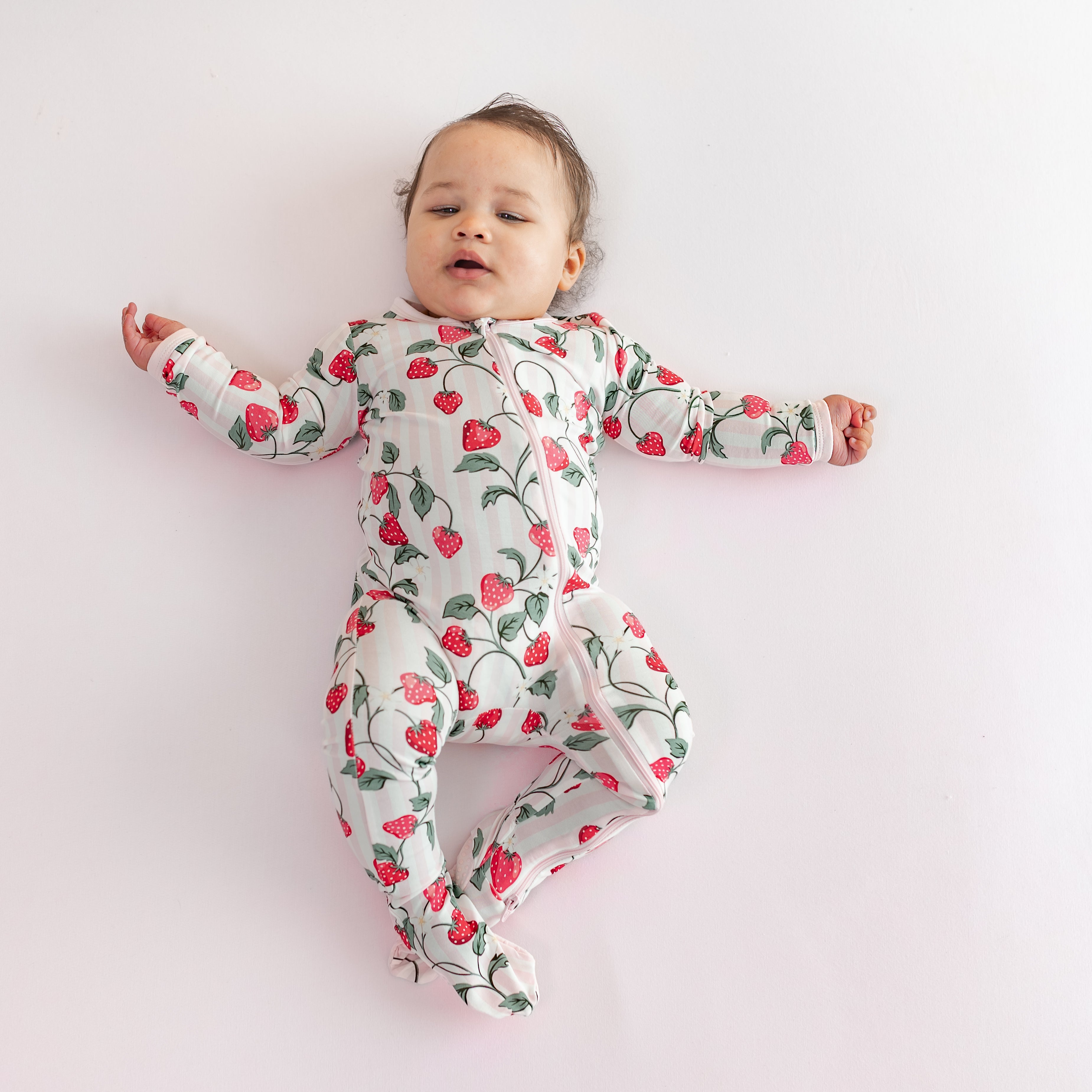 Young infant wearing the Zippered Footie in Strawberry Fields laying on a light pink surface
