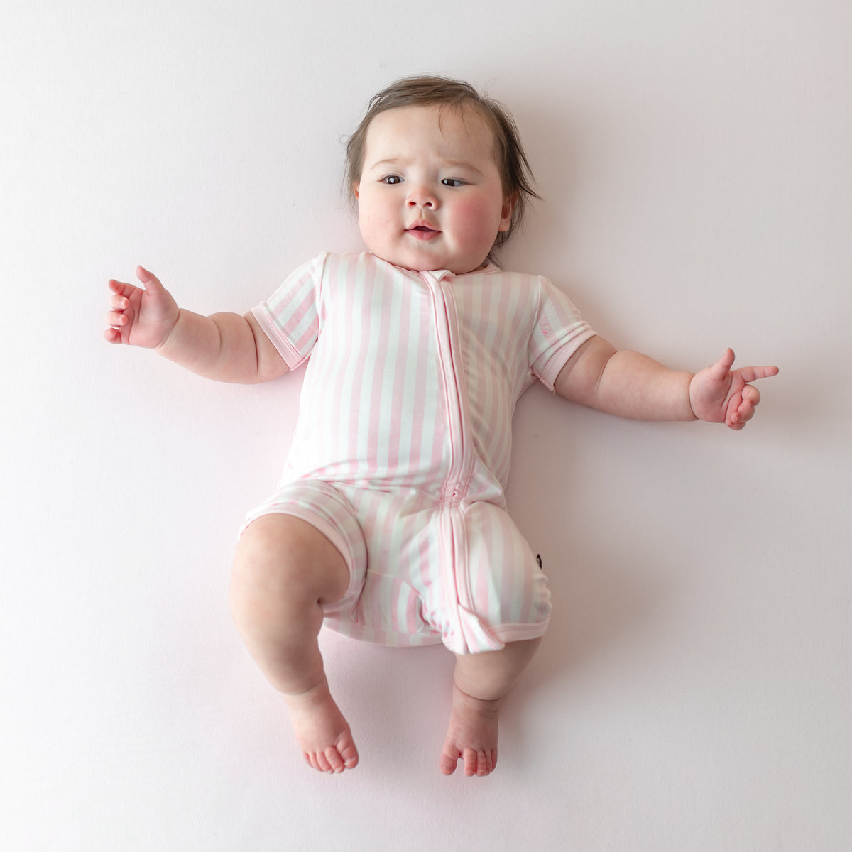 Infant laying on a light pink surface wearing the buttery soft Zipper Shortall Romper in Small Sakura Stripe