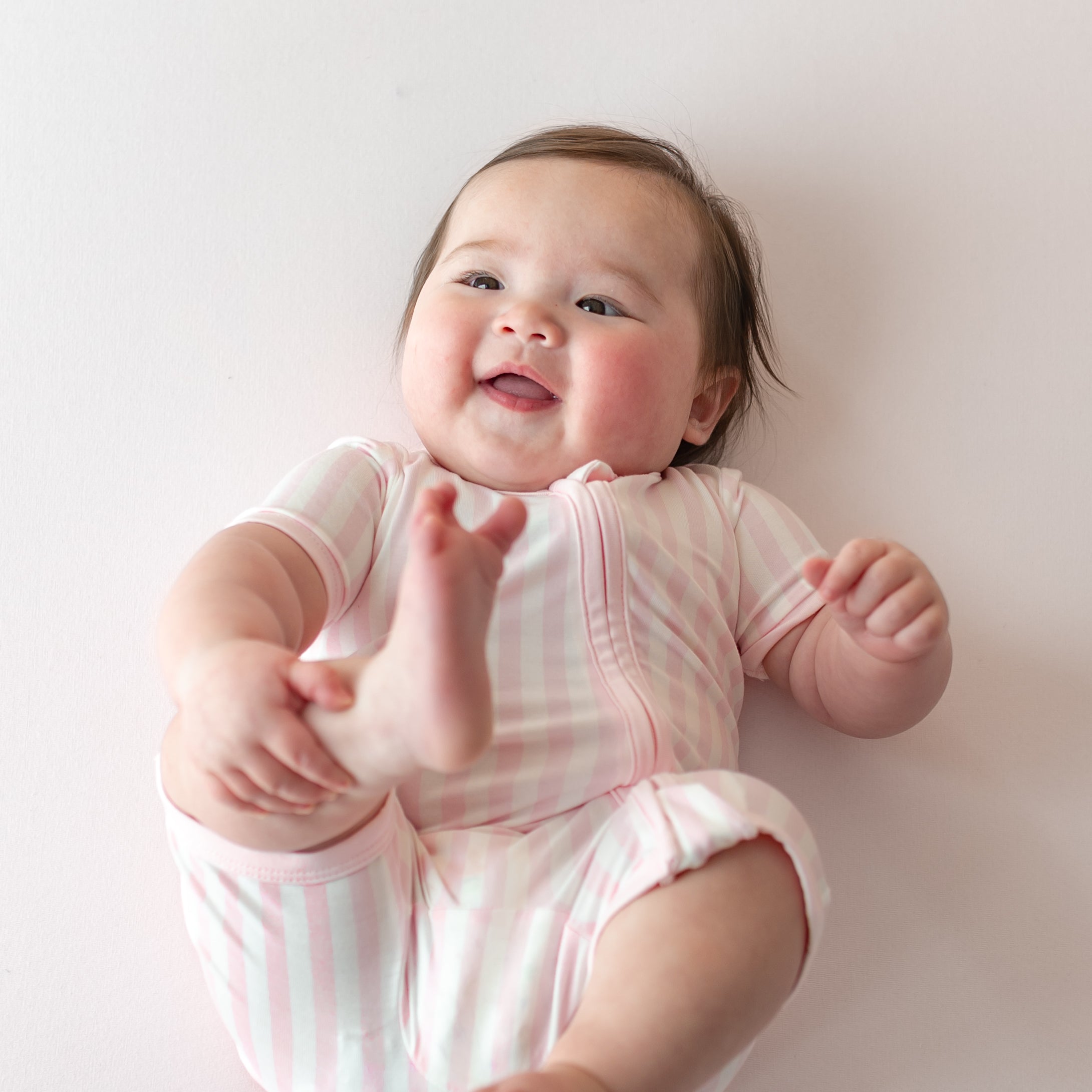 Smiling infant laying on her back on a pink surface wearing the Zipper Shortall Romper in Small Sakura Stripe