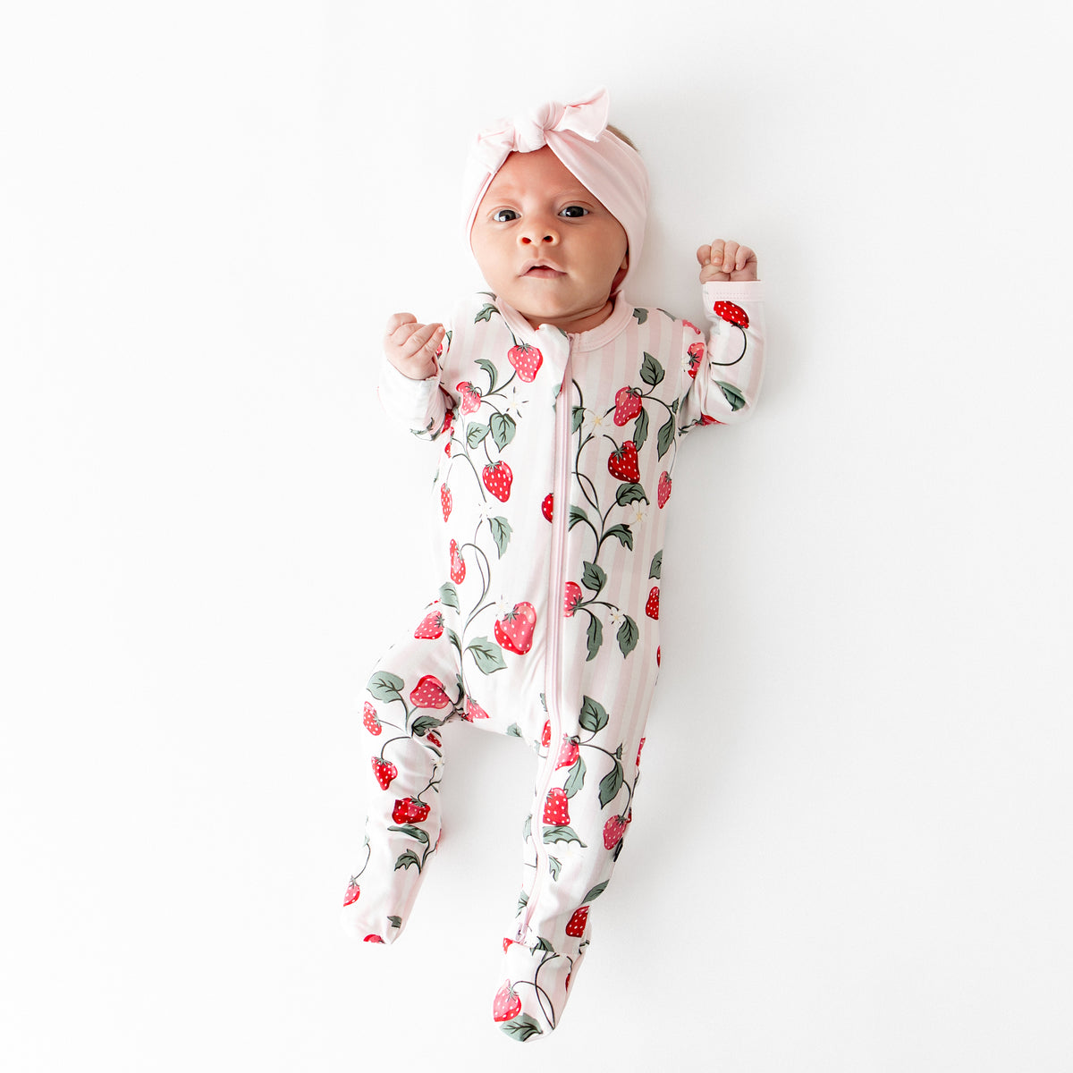 Newborn laying on a light neutral surface wearing the Zippered Footie in Strawberry Fields with a pink bow