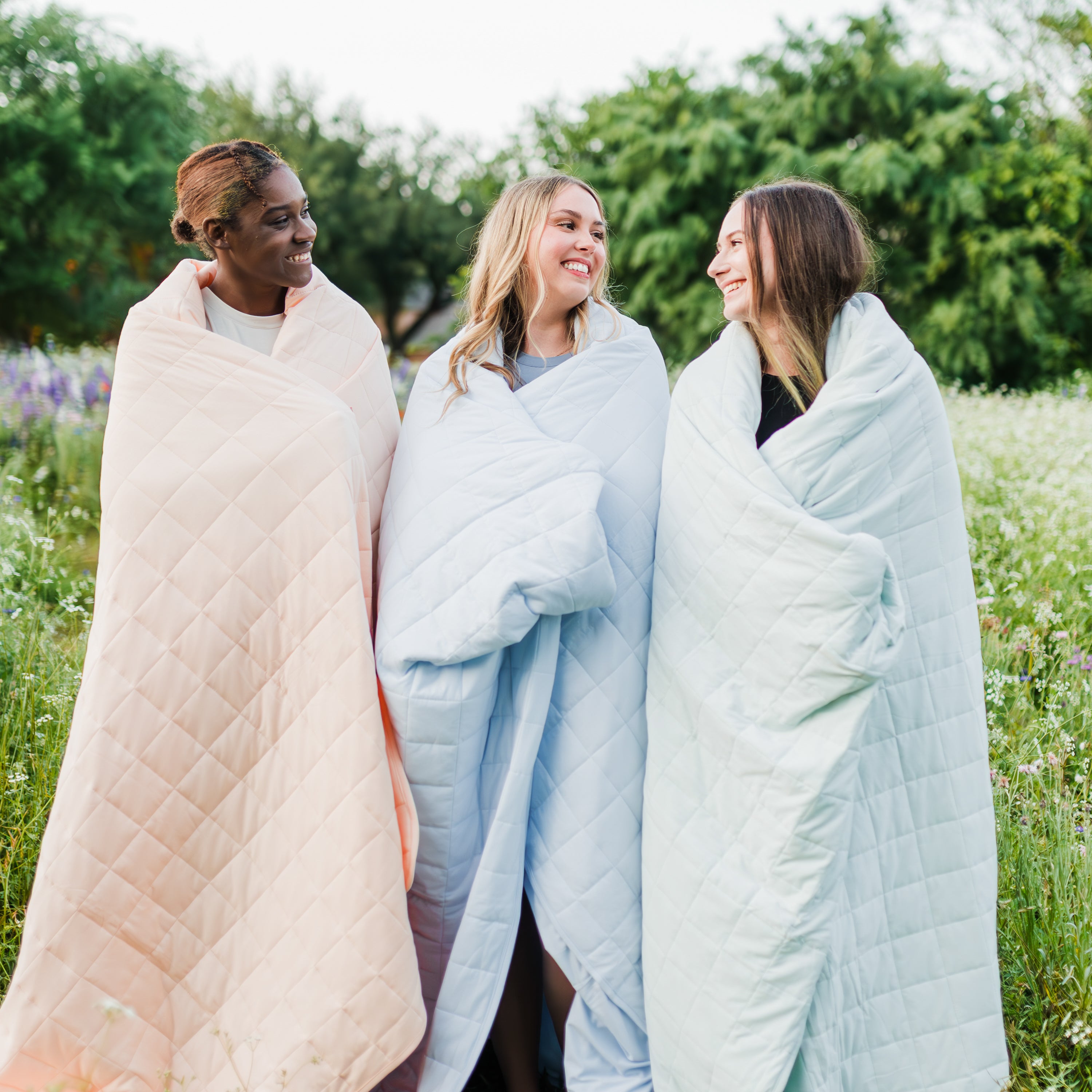 3 women wrapped in Kyte Baby Adult Blankets in the colors Canyon, Mist and Dew in a wildflower field