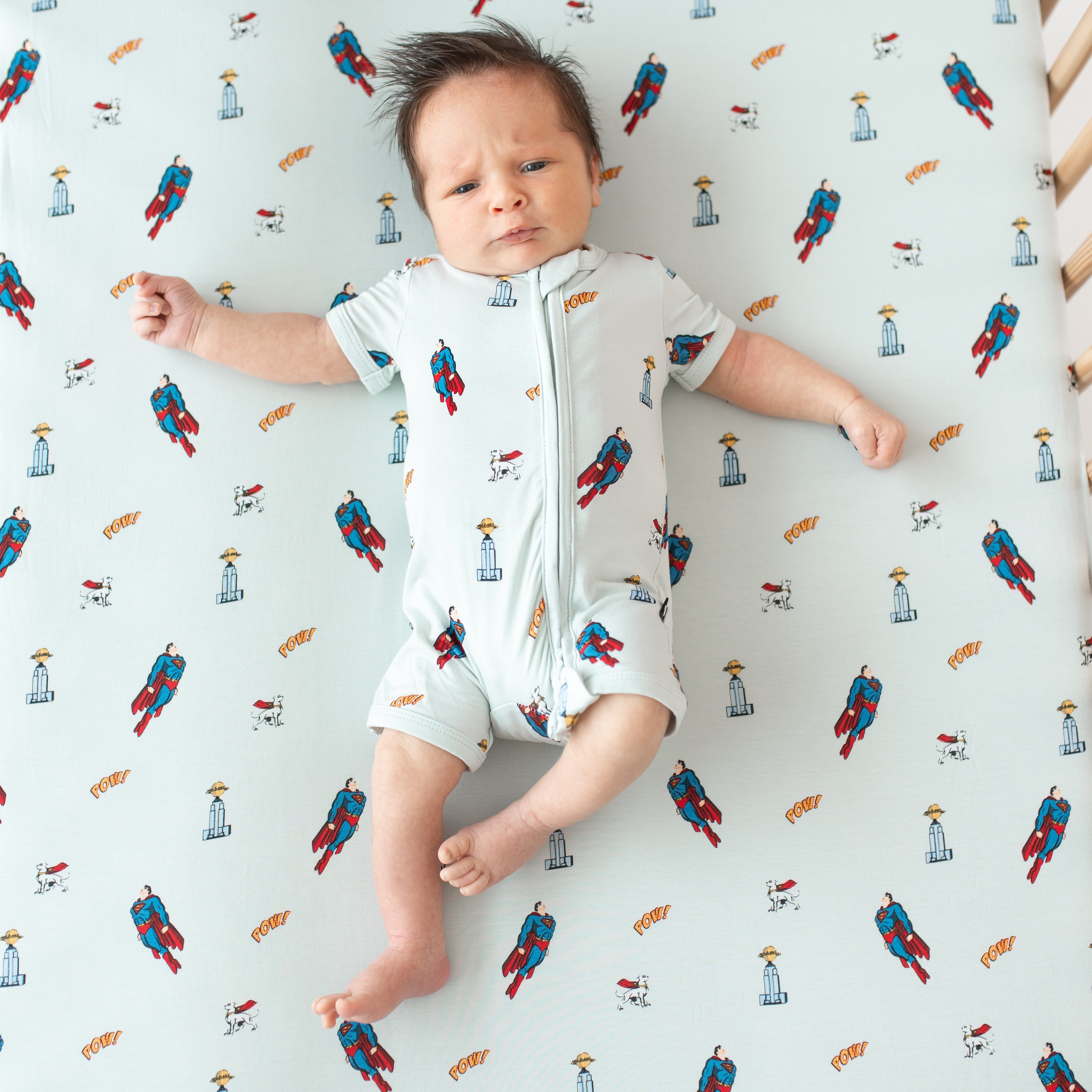 Newborn laying in a crib on a Crib Sheet in Superman™ wearing the matching Zippered Shortall Romper