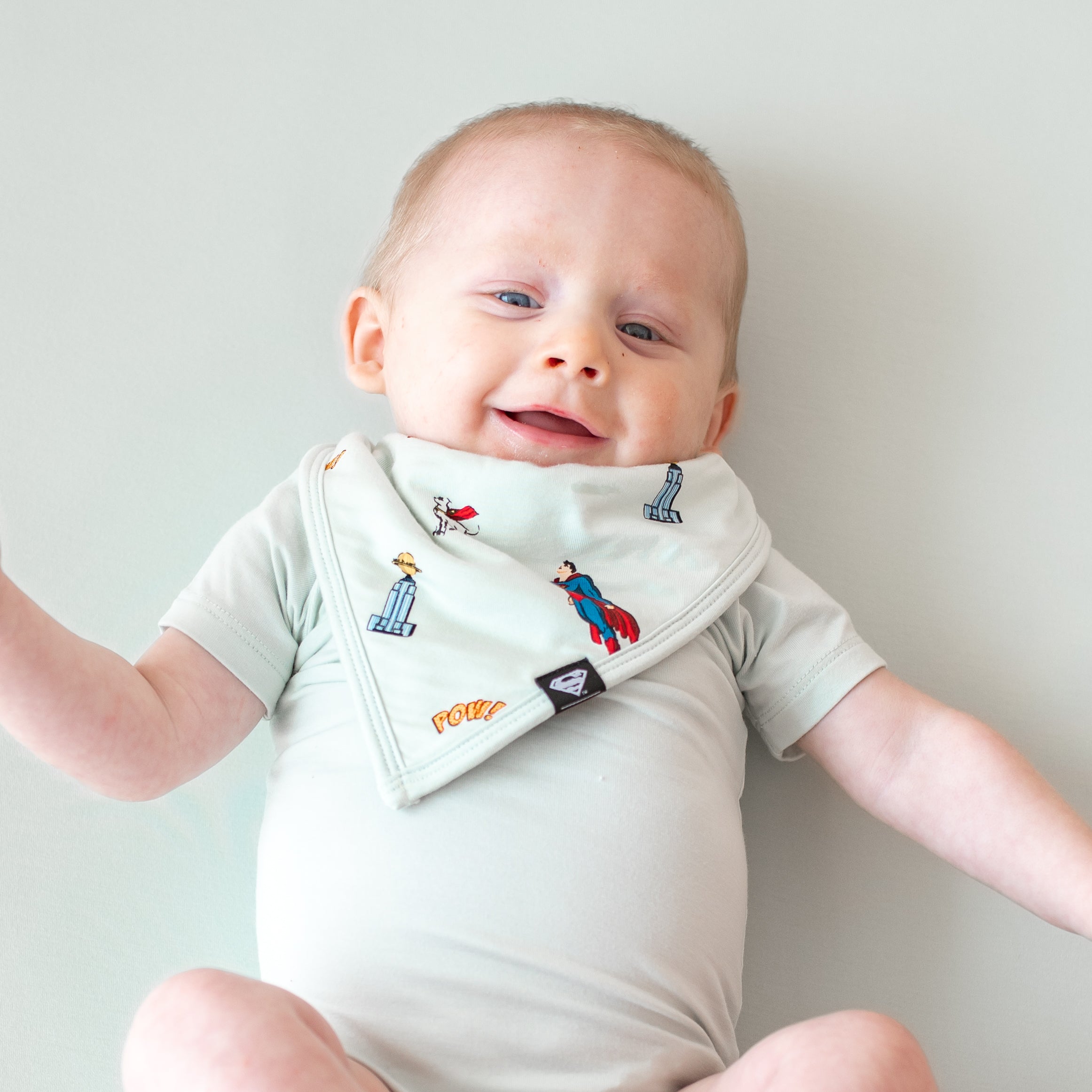 Smiling infant wearing the Bib in Superman™ paired with a Dew Short Sleeve Bodysuit