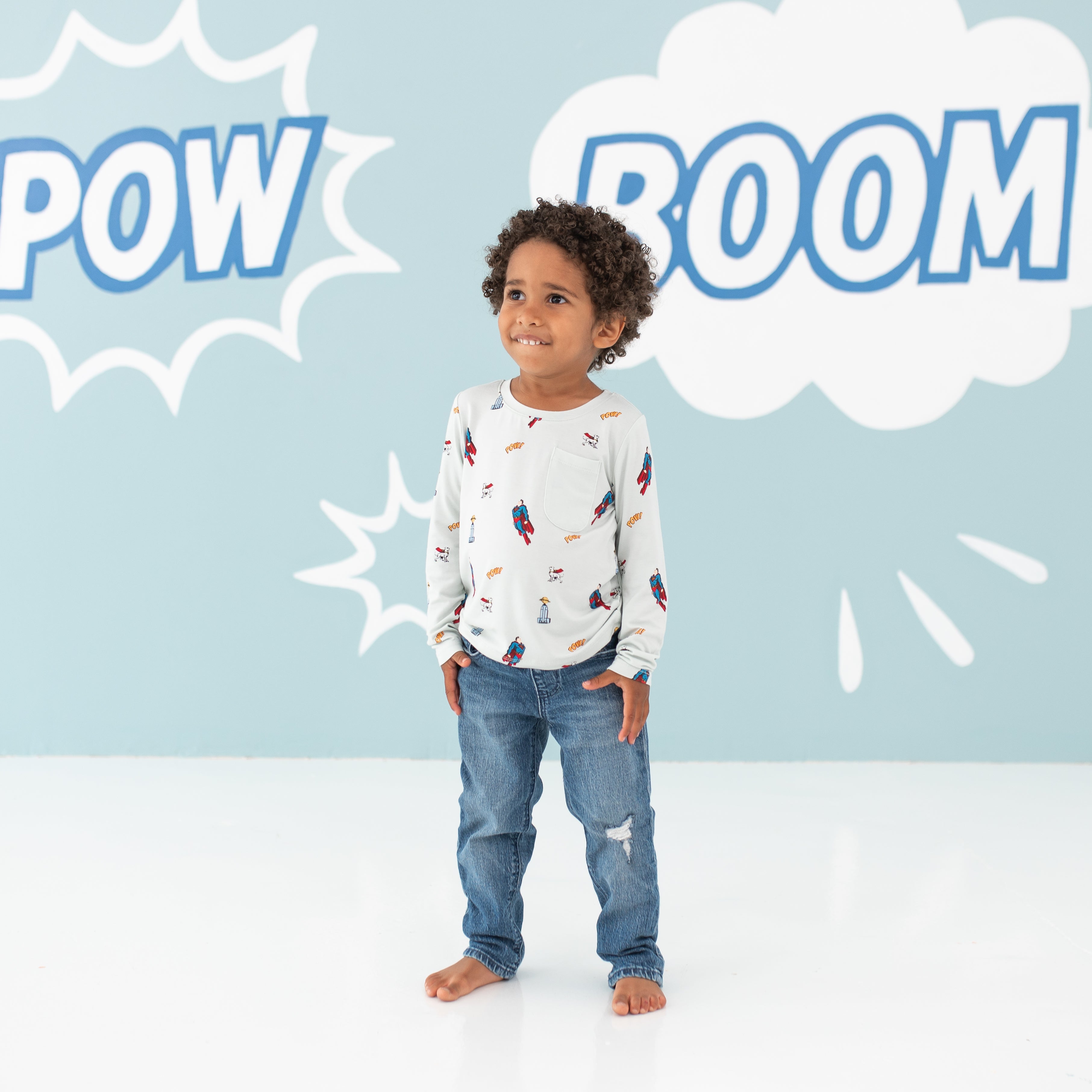 Smiling model wearing the Long Sleeve Toddler Crew Neck Tee in Superman™ paired with jeans