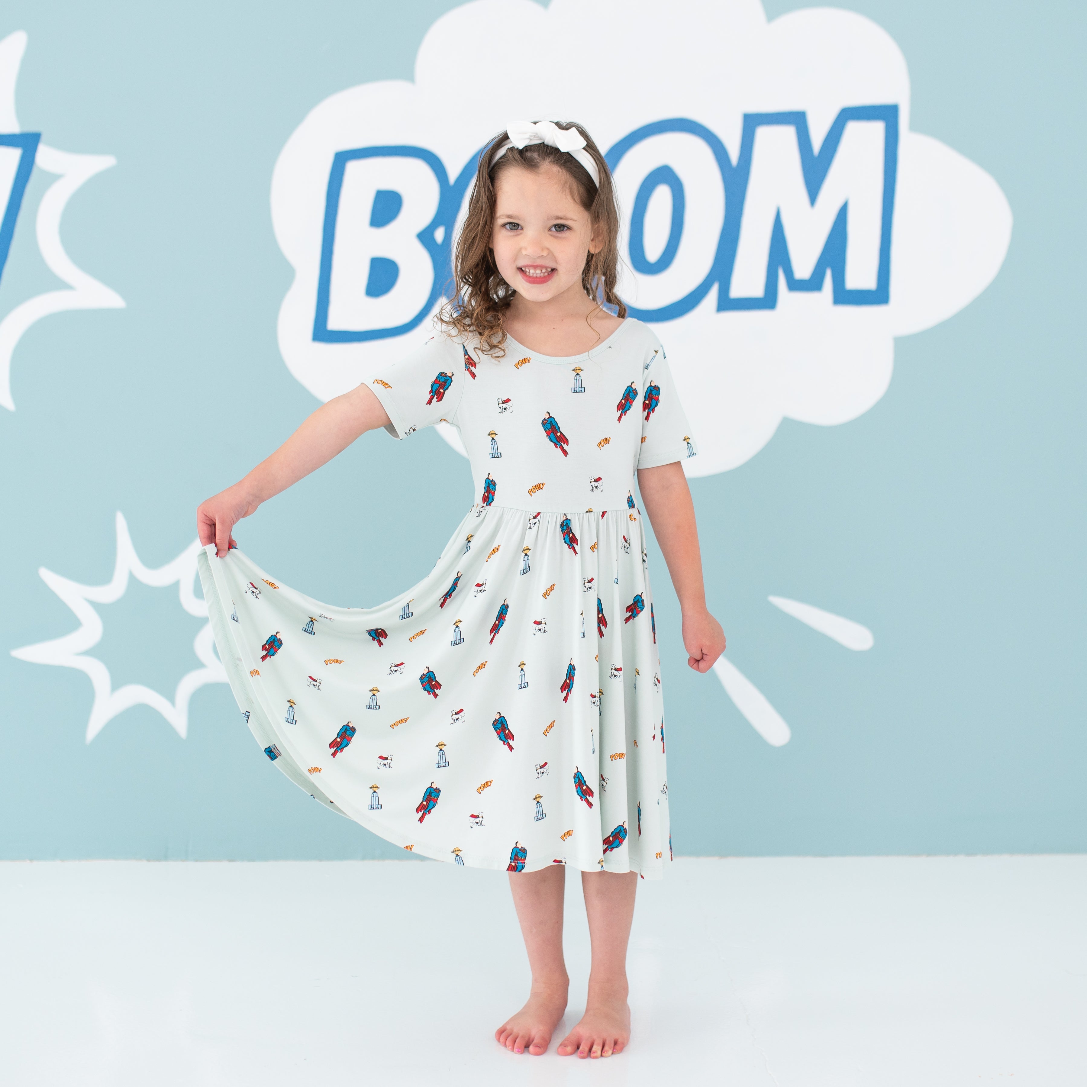 Young girl standing holding one side of her Twirl Dress in Superman™