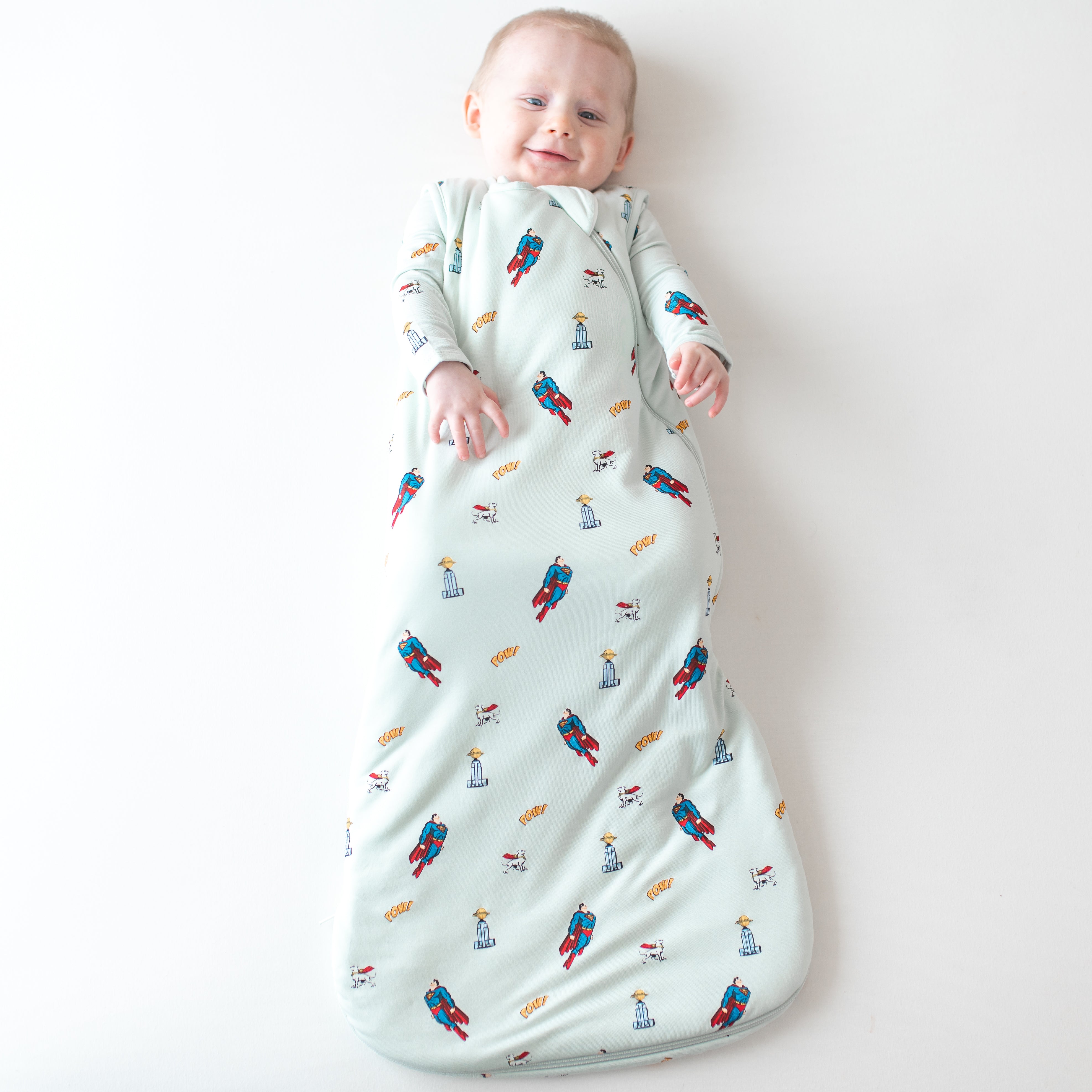 Smiling infant wearing the Sleep Bag in Superman™ 1.0 paired with a matching long sleeve romper