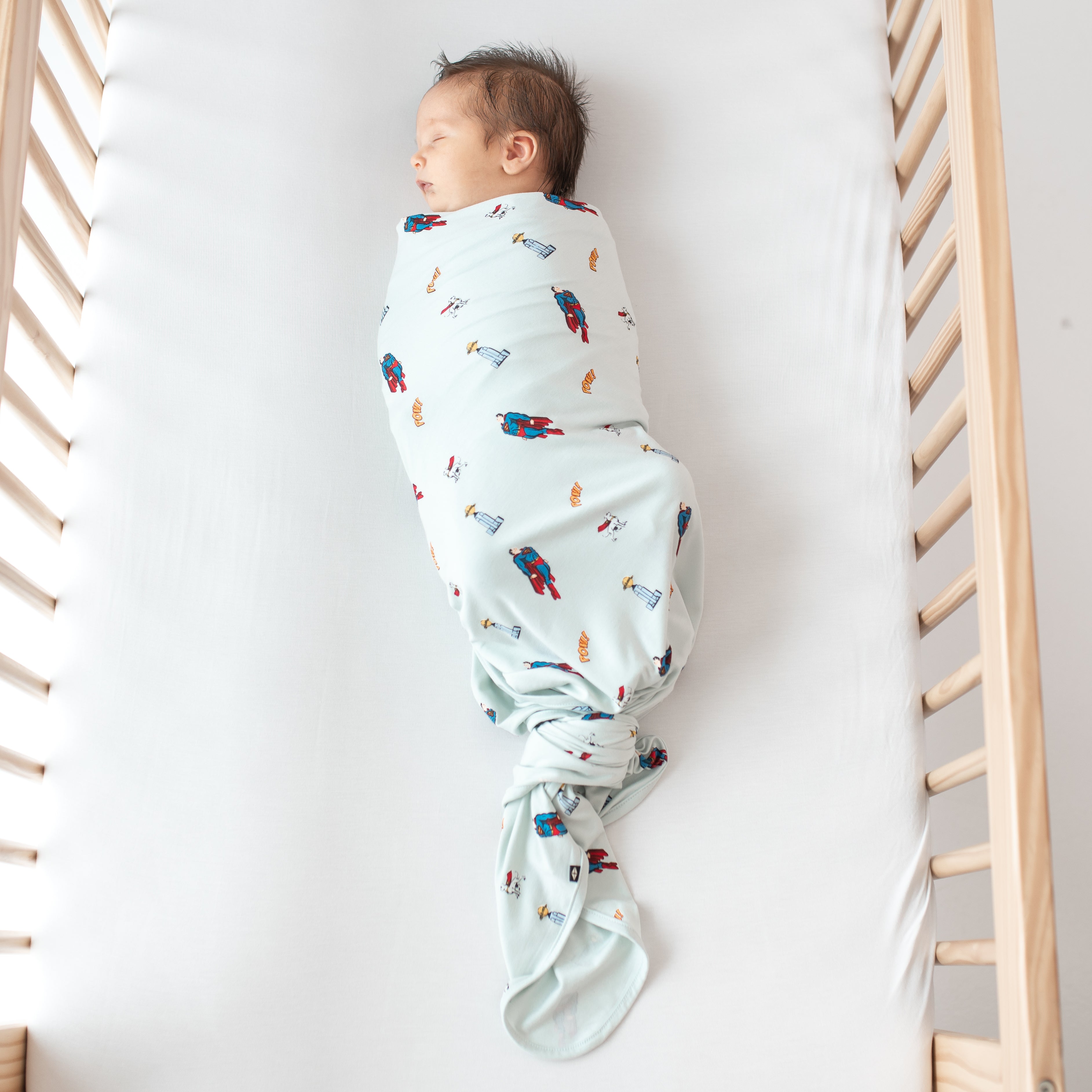 Sleeping newborn laying in a crib swaddled in the Swaddle Blanket in Superman™