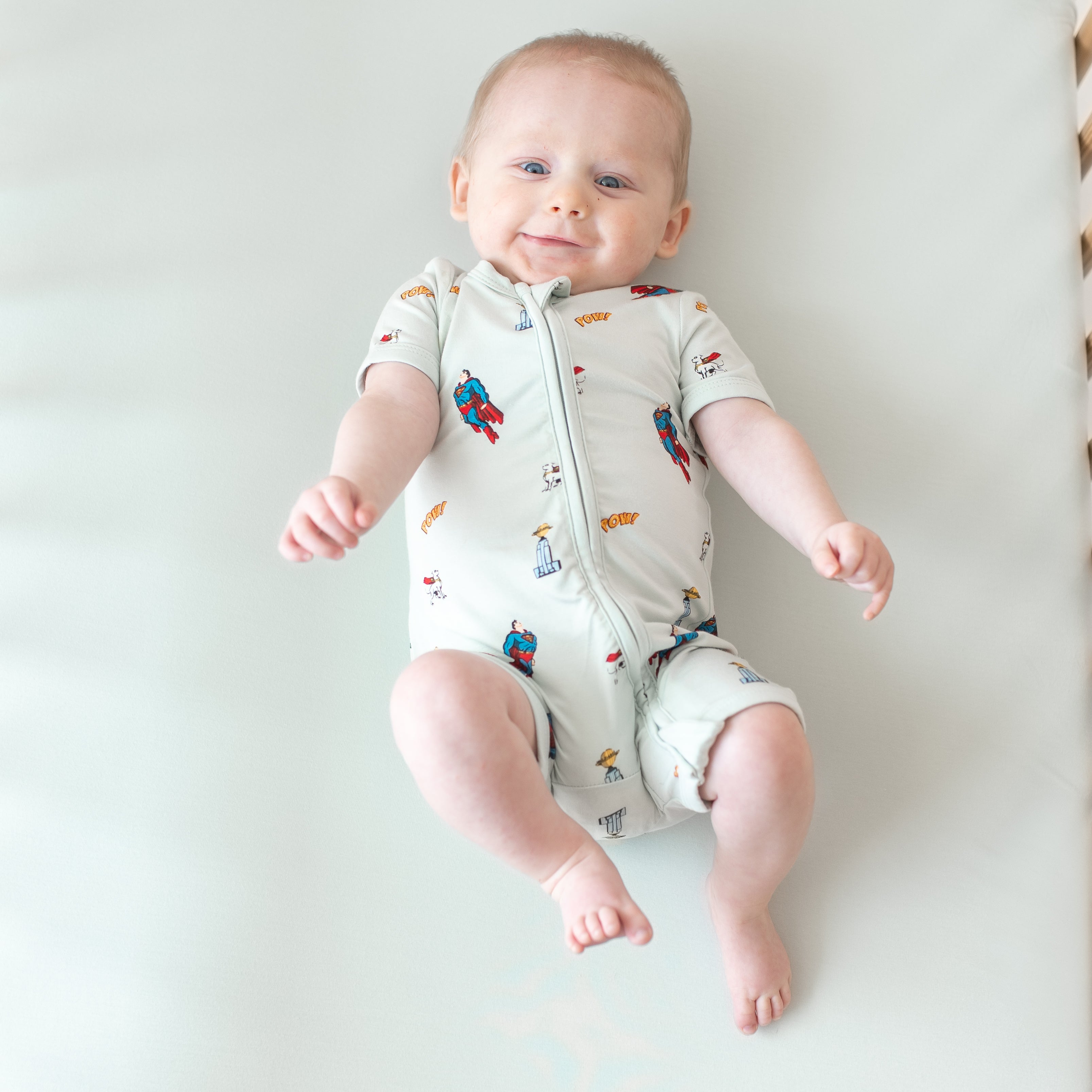 Smiling infant laying in a crib wearing the Zipper Shortall Romper in Superman™