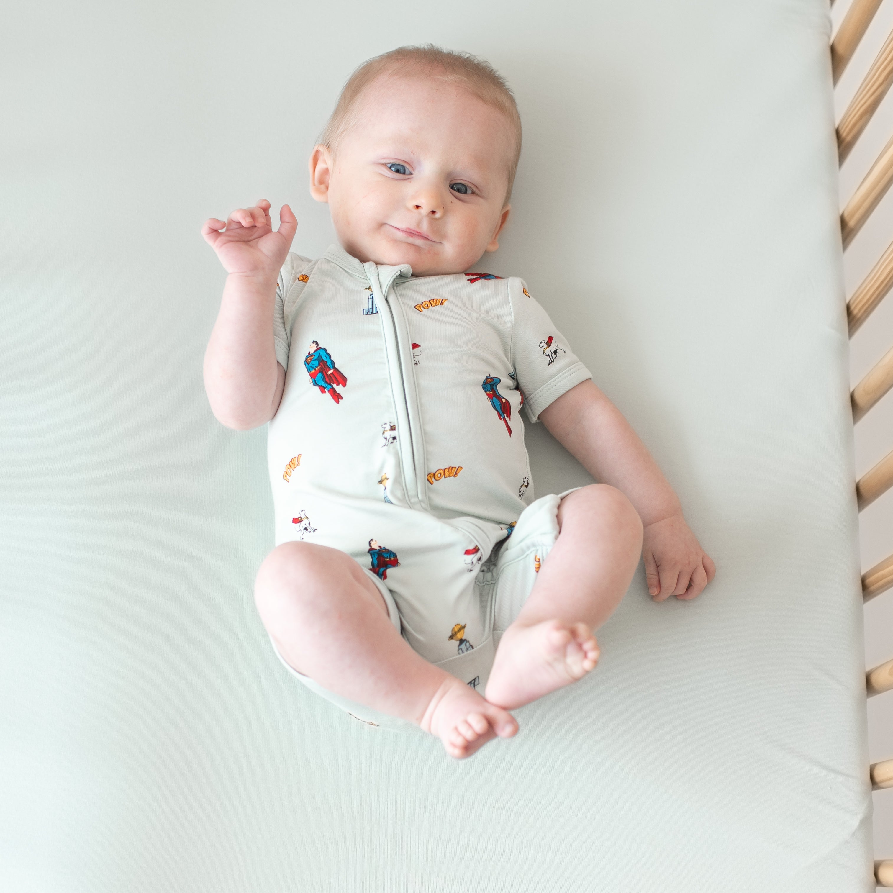 Smiling infant laying in a crib with his feet in the air wearing the Zipper Shortall Romper in Superman™