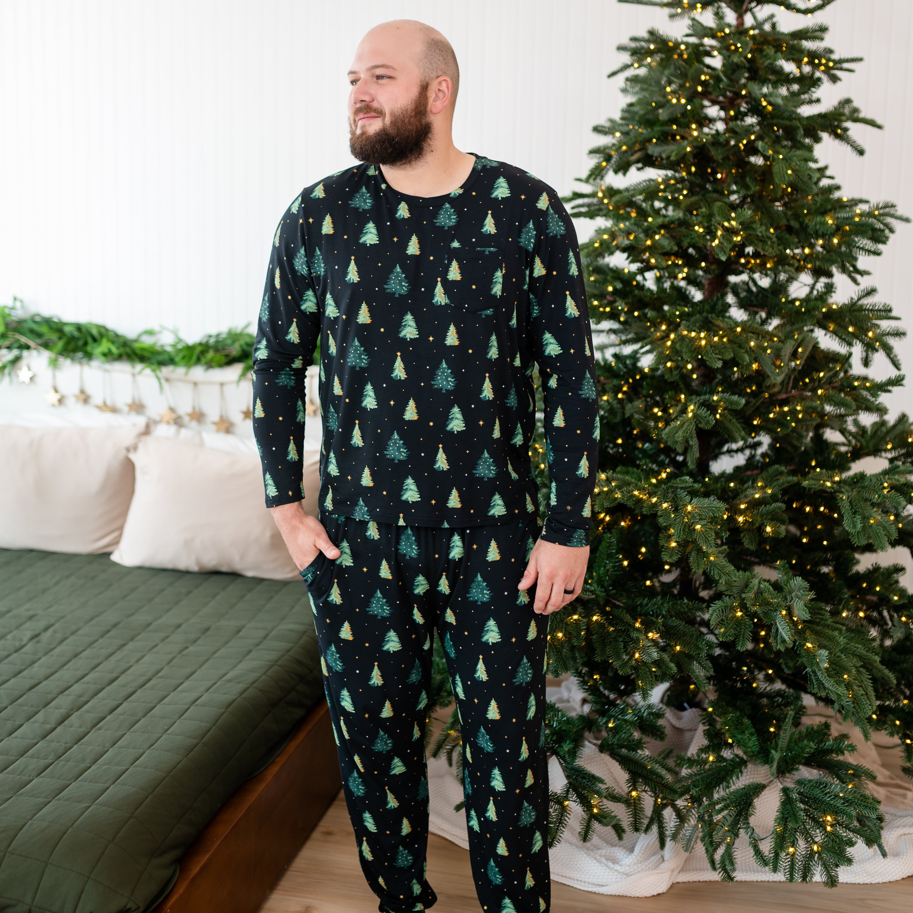Male model standing with one hand in the pocket of the pants of the Men's Jogger Set in Twinkle Tree standing in front of a bed and a christmas tree