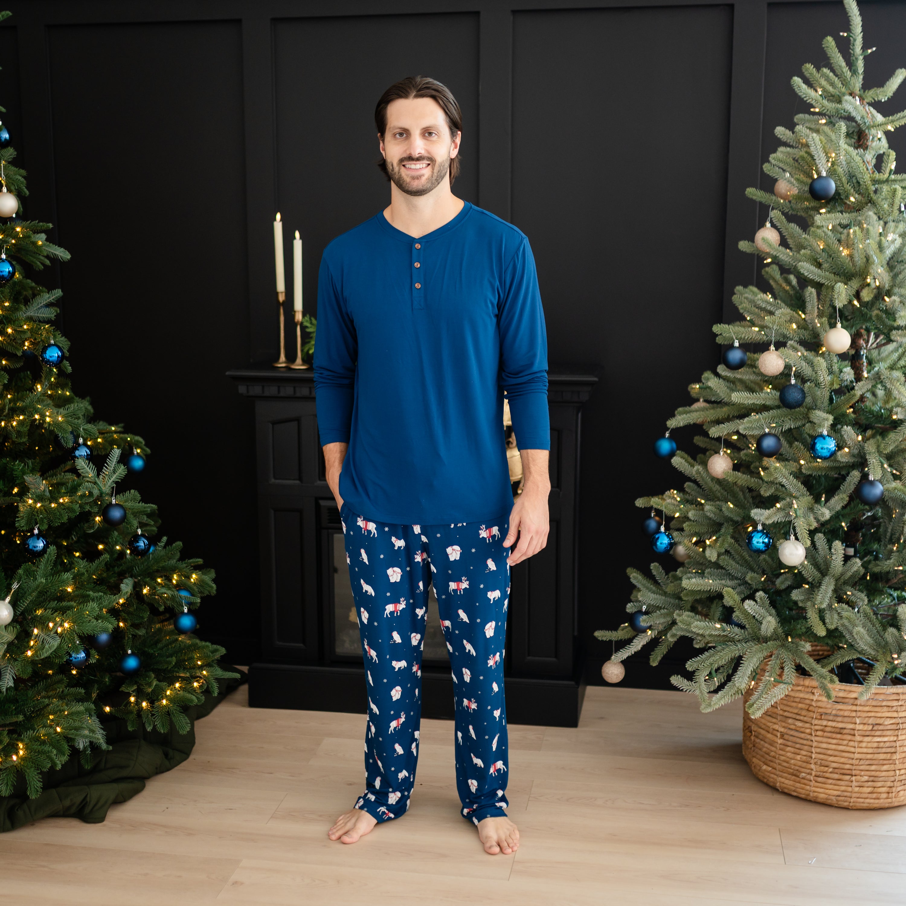 Smiling male model standing between two decorated christmas trees wearing the Men's Long Sleeve Henley Top in Tahoe and the Men's Lounge Pant in Frosty Friends