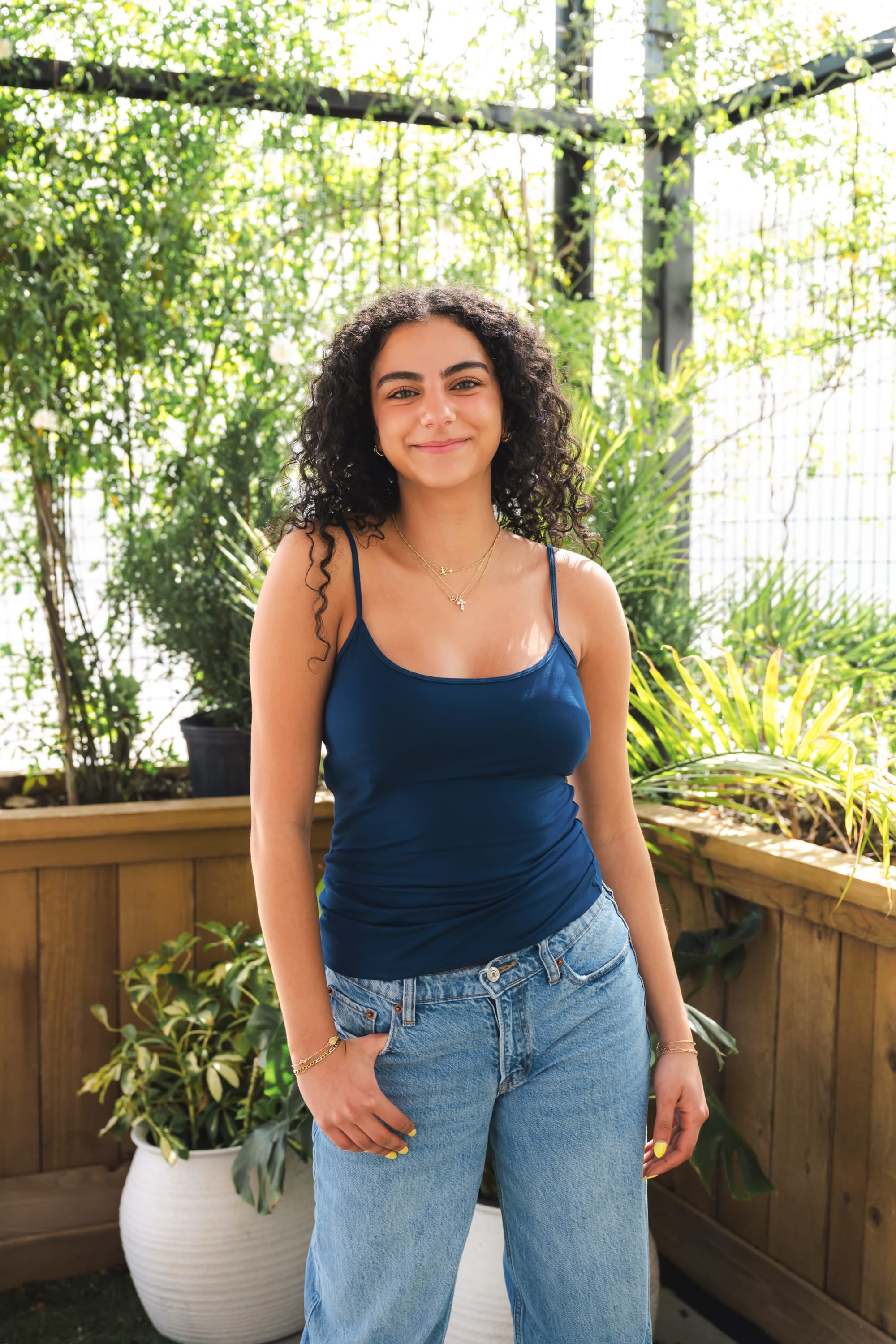 Woman wearing a blue tank top and jeans standing outdoors with plants in the background