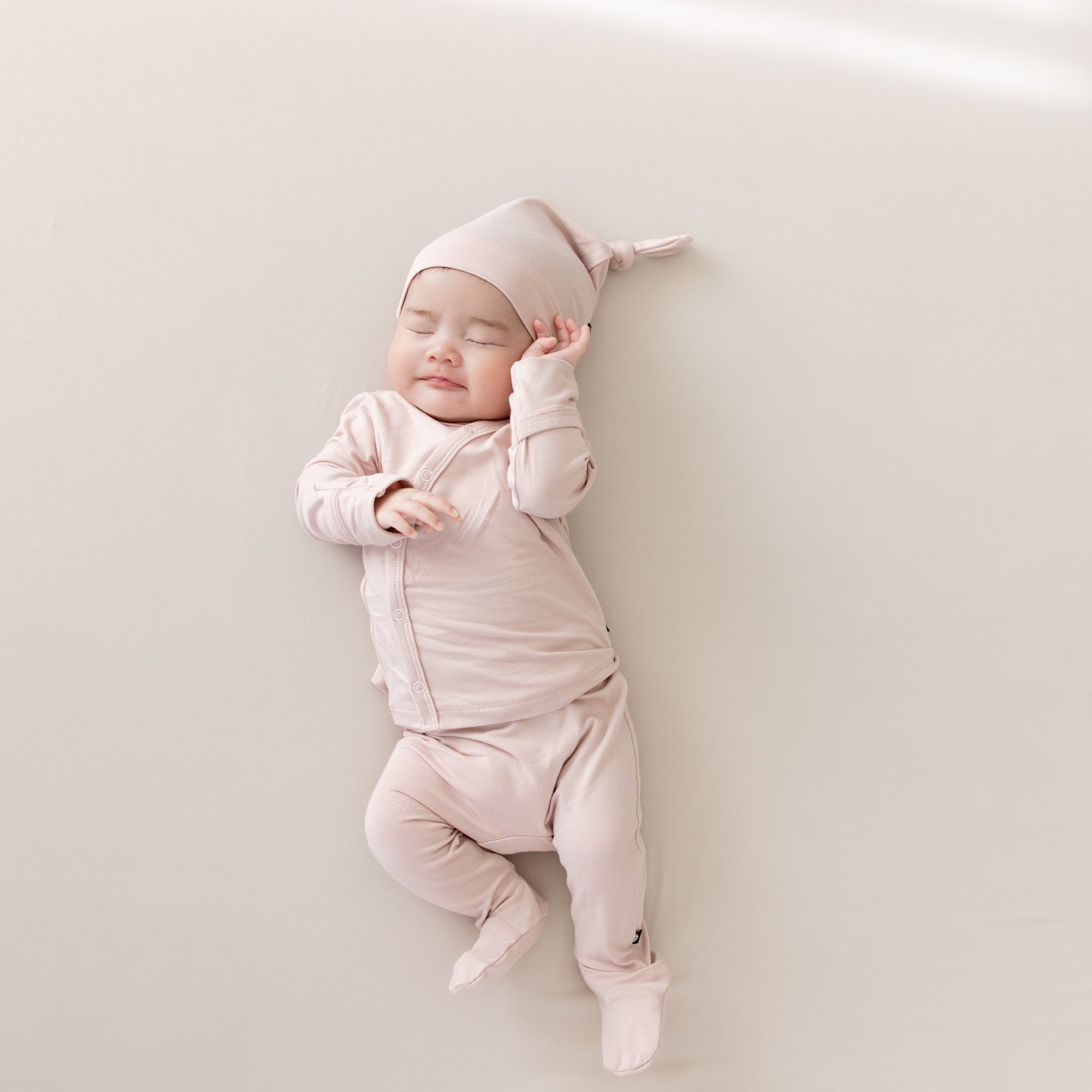 Baby laying on an Oat surface while wearing the Bamboo Take Me Home Set with Cap in Blush