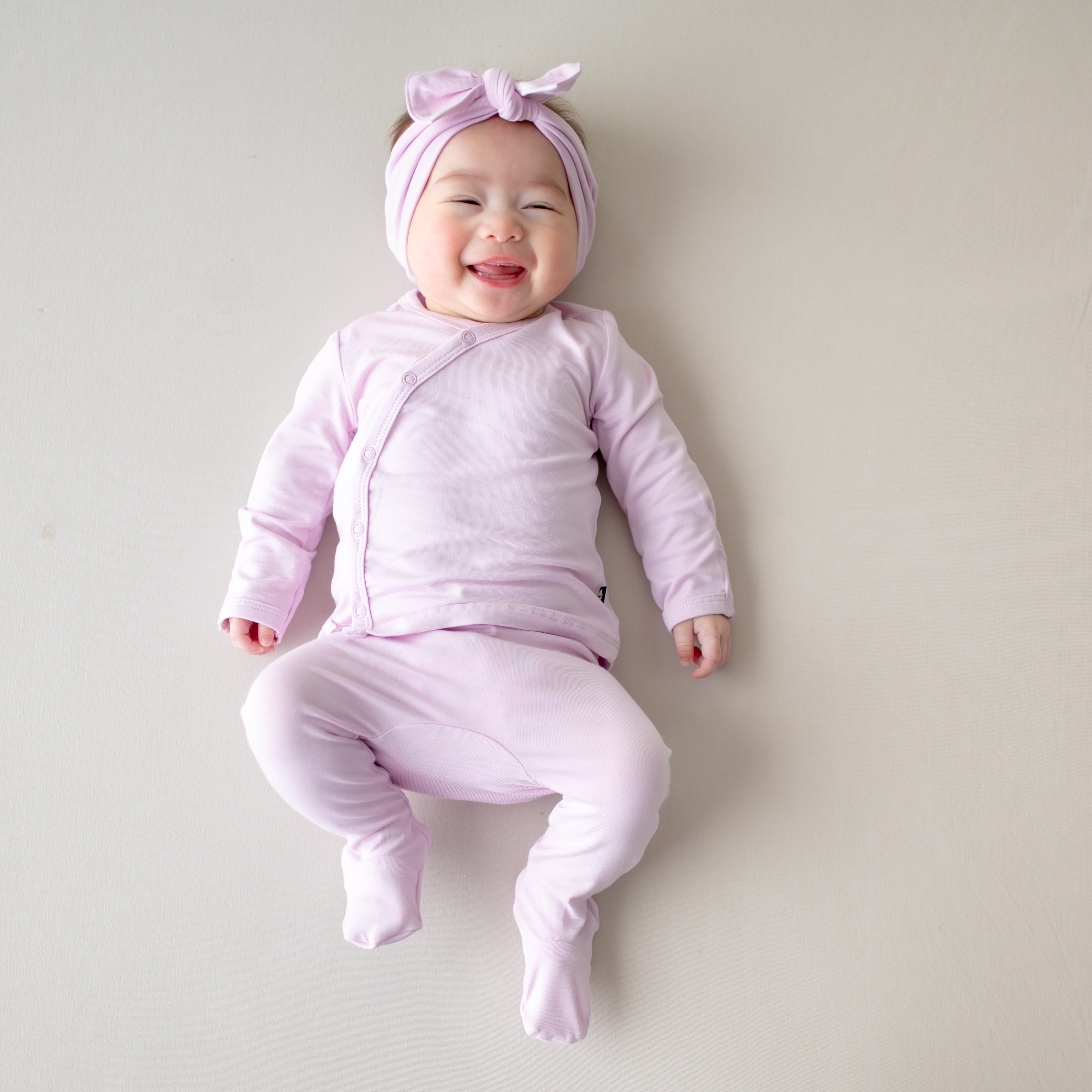Smiling newborn laying on a light neutral surface wearing the Take Me Home Set with Bow in Thistle