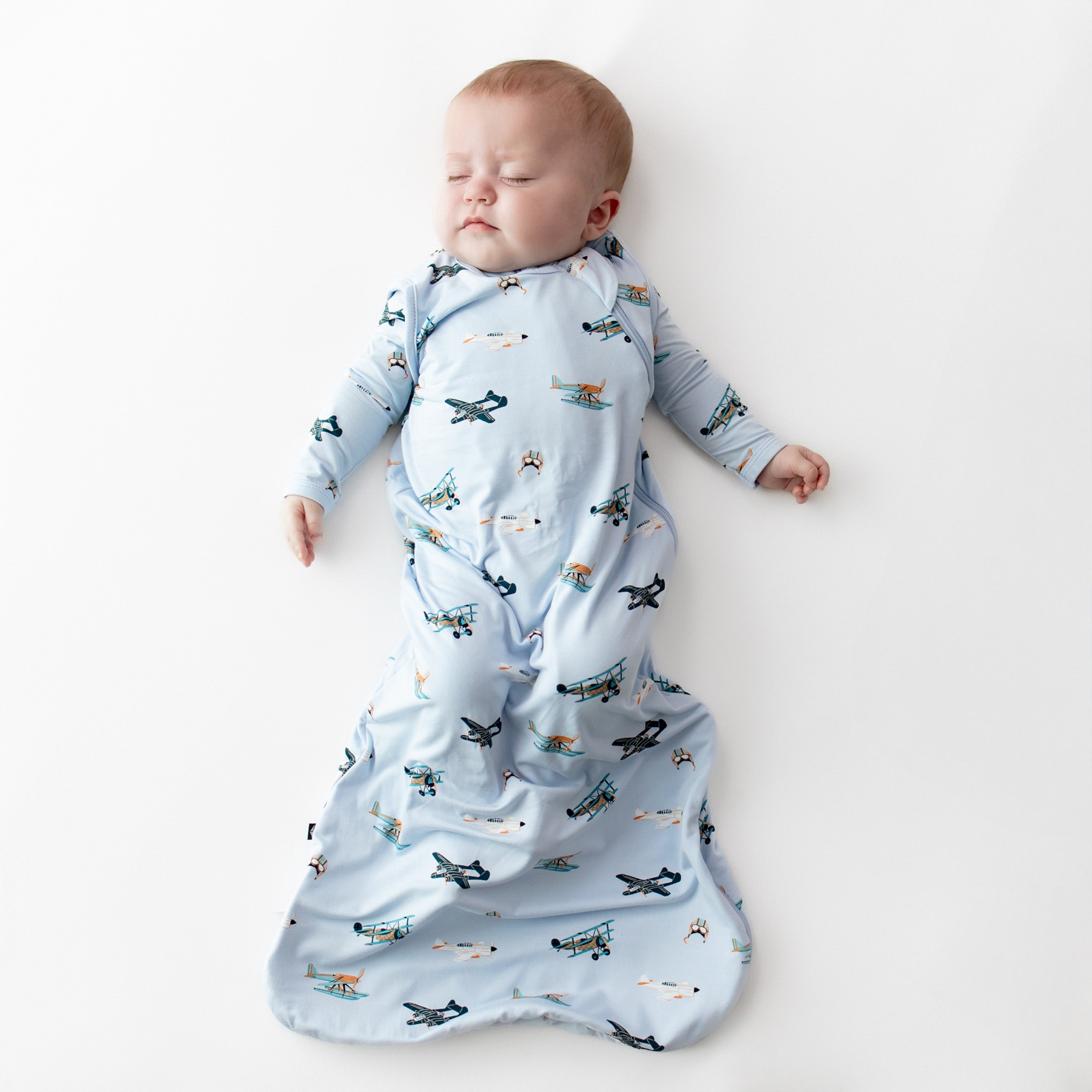 Sleeping infant laying on a white surface wearing the Sleep Bag in Take Off 0.5 wearing a matching long sleeve romper