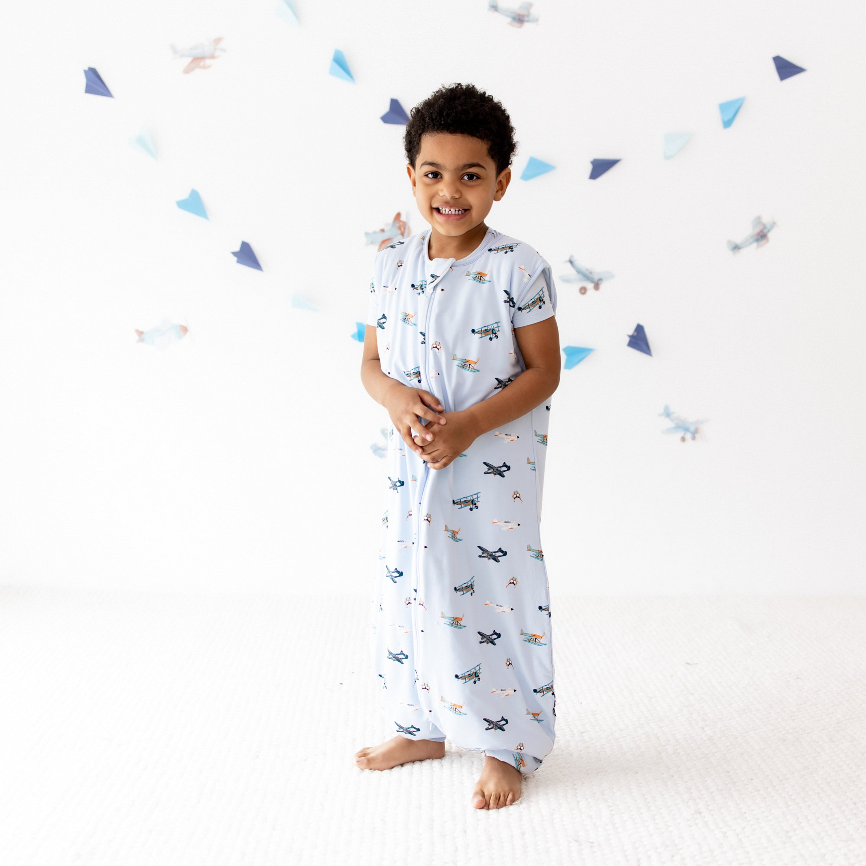 Smiling boy standing on a white carpet wearing the Sleep Bag Walker in Take Off 1.0