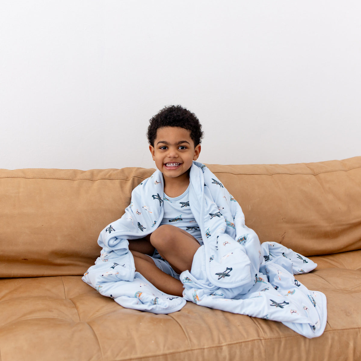 Young boy sitting on a couch with the Toddler Blanket in Take Off 1.0 around his shoulders