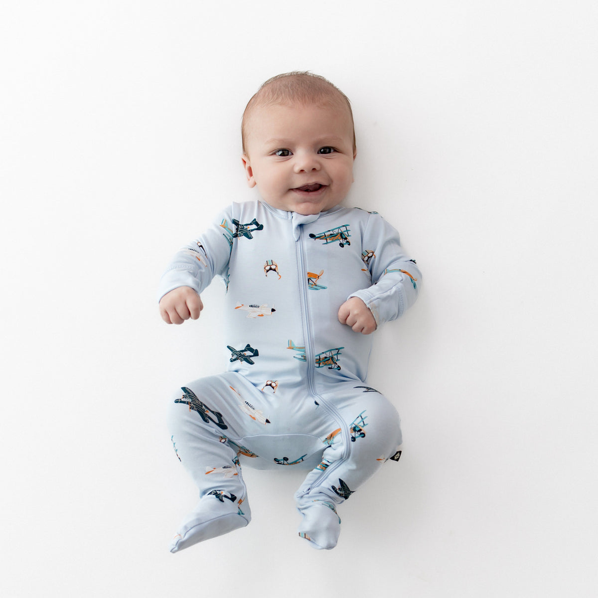 Infant laying on a white surface wearing the Zippered Footie in Take Off